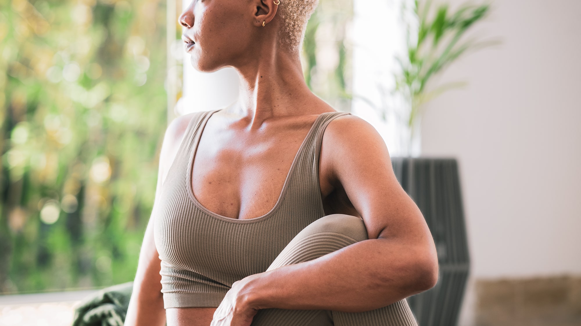 Woman in beige yoga outfit doing seated twist on mat, looking out window.