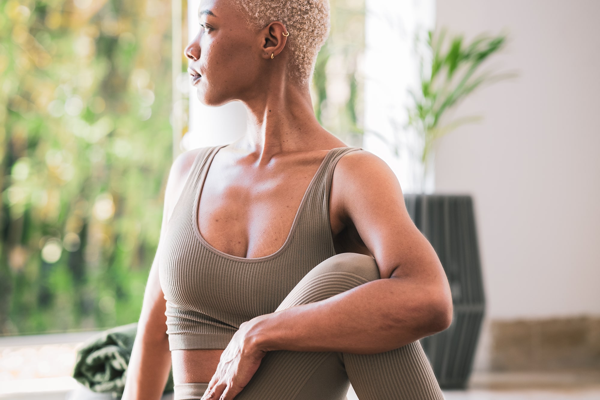 Woman in beige yoga outfit doing seated twist on mat, looking out window.