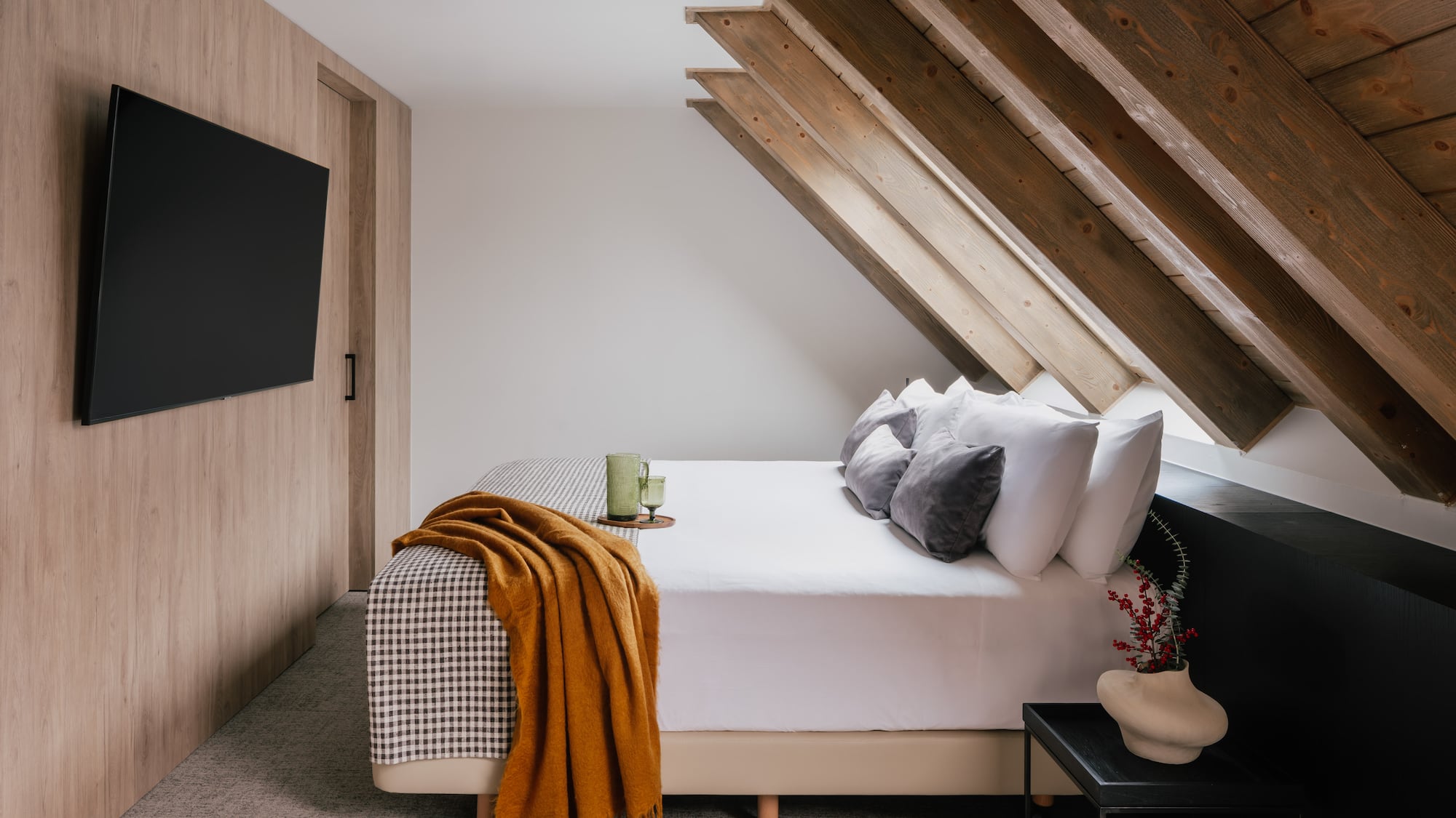 Modern bedroom with slanted wooden ceiling, white bed, orange throw, and TV.