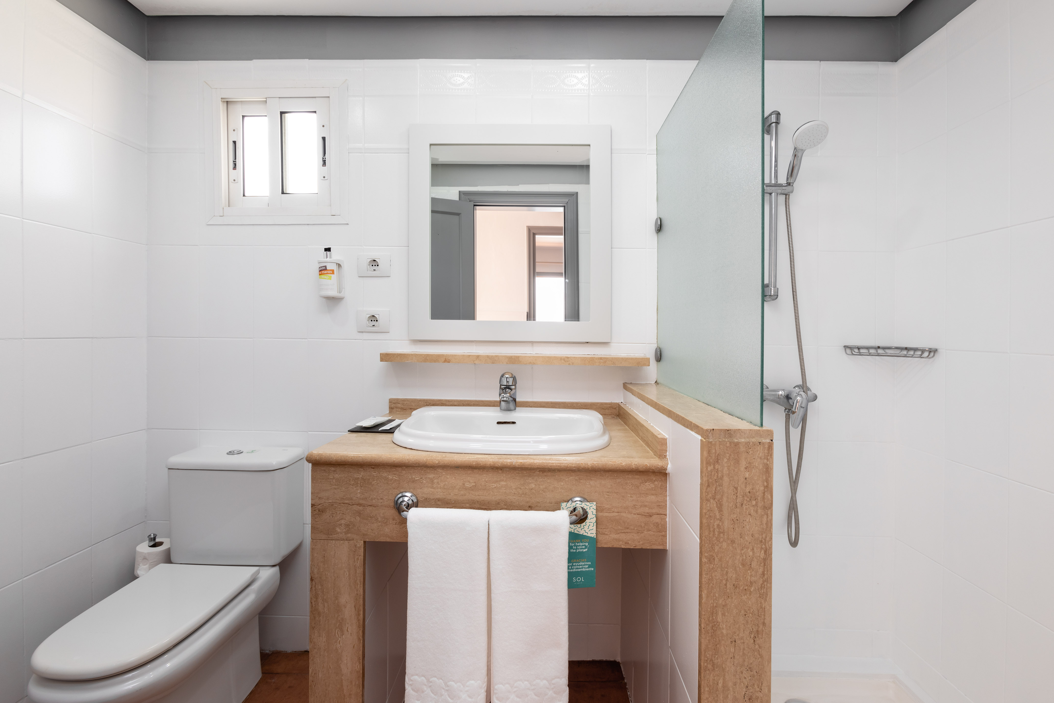 White tiled bathroom with vanity sink, mirror, toilet, and walk-in shower.