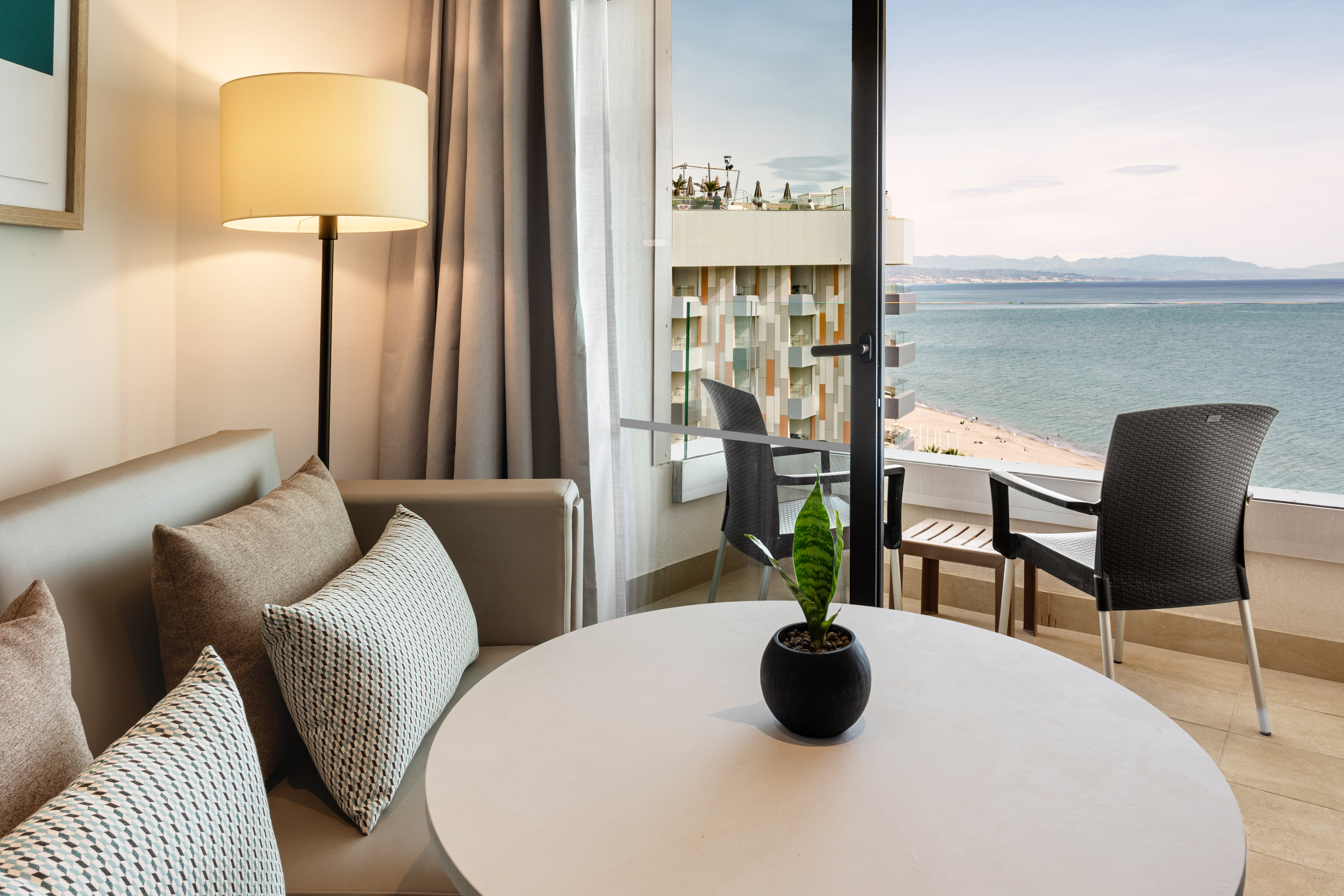 Modern hotel suite with ocean view balcony, sandy beach, and distant mountains.