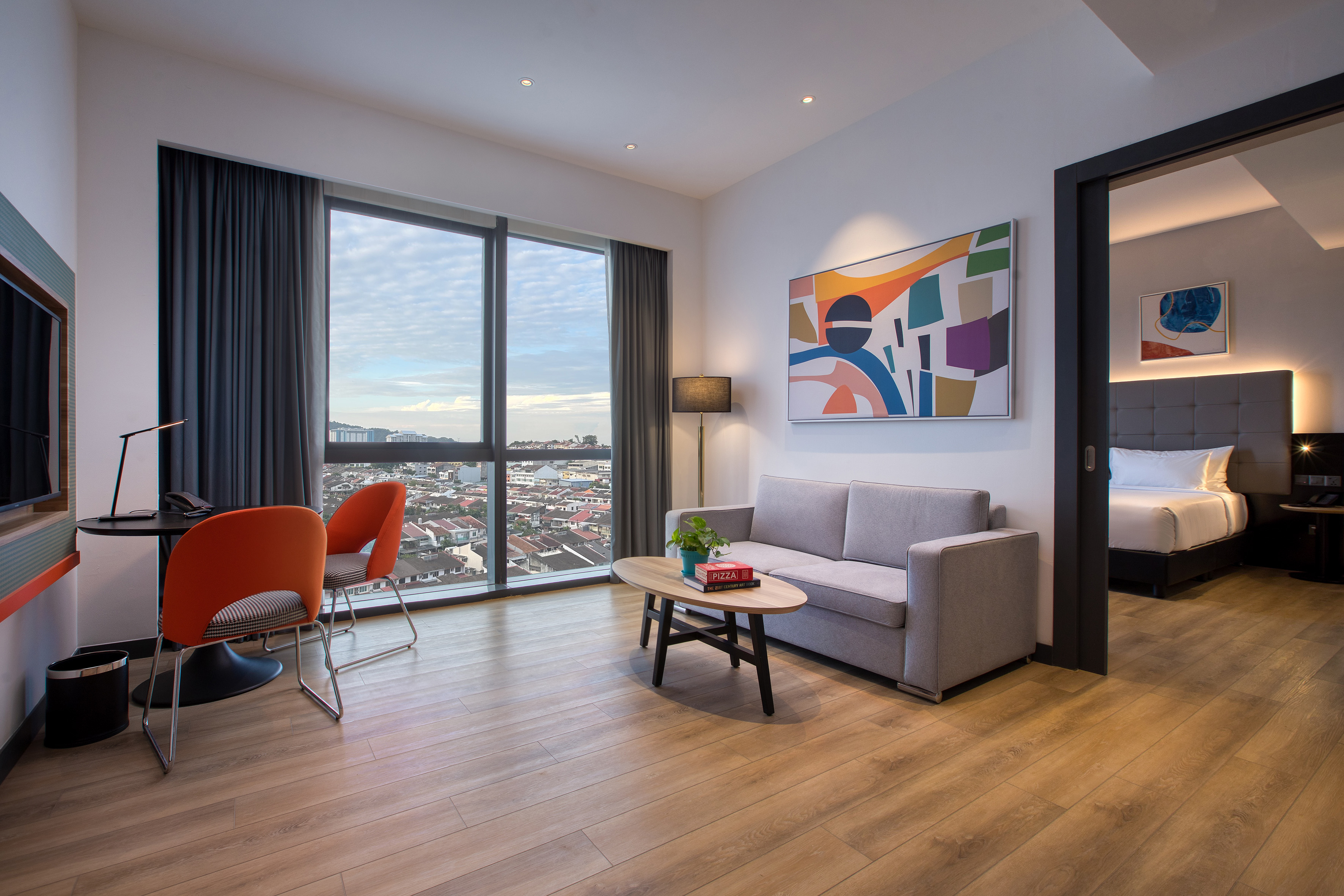 Modern hotel suite living area with city view, sofa, desk, and adjoining bedroom.