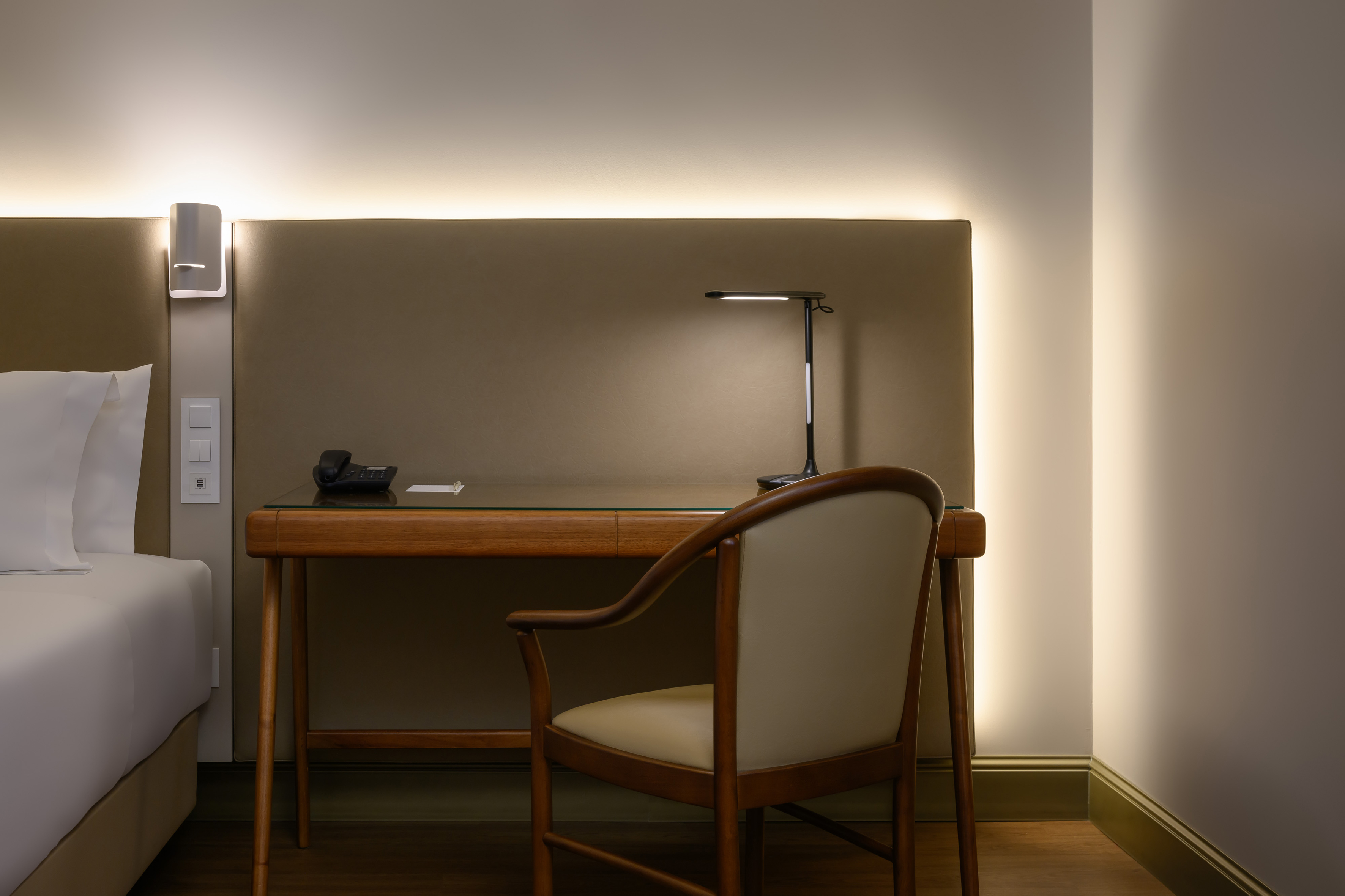 Hotel room desk with sleek lamp, telephone, and elegant chair beside bed.