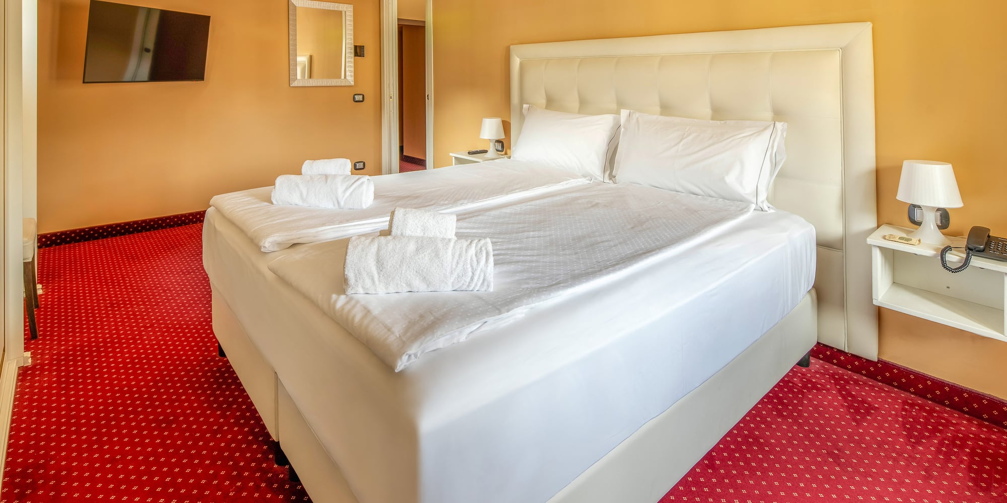 Hotel room with queen bed, white linen, red carpet, and orange walls.