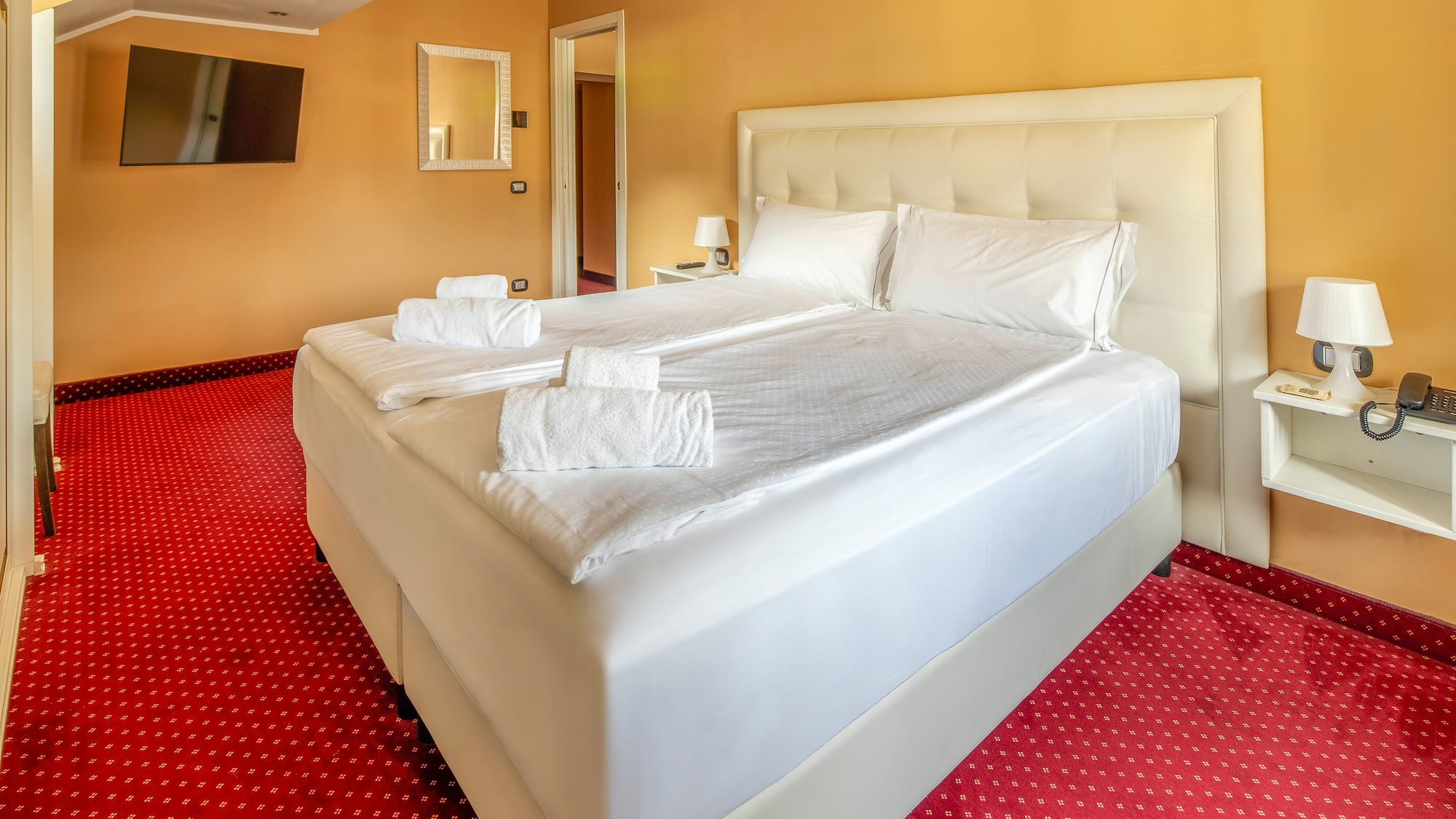 Hotel room with queen bed, white linen, red carpet, and orange walls.