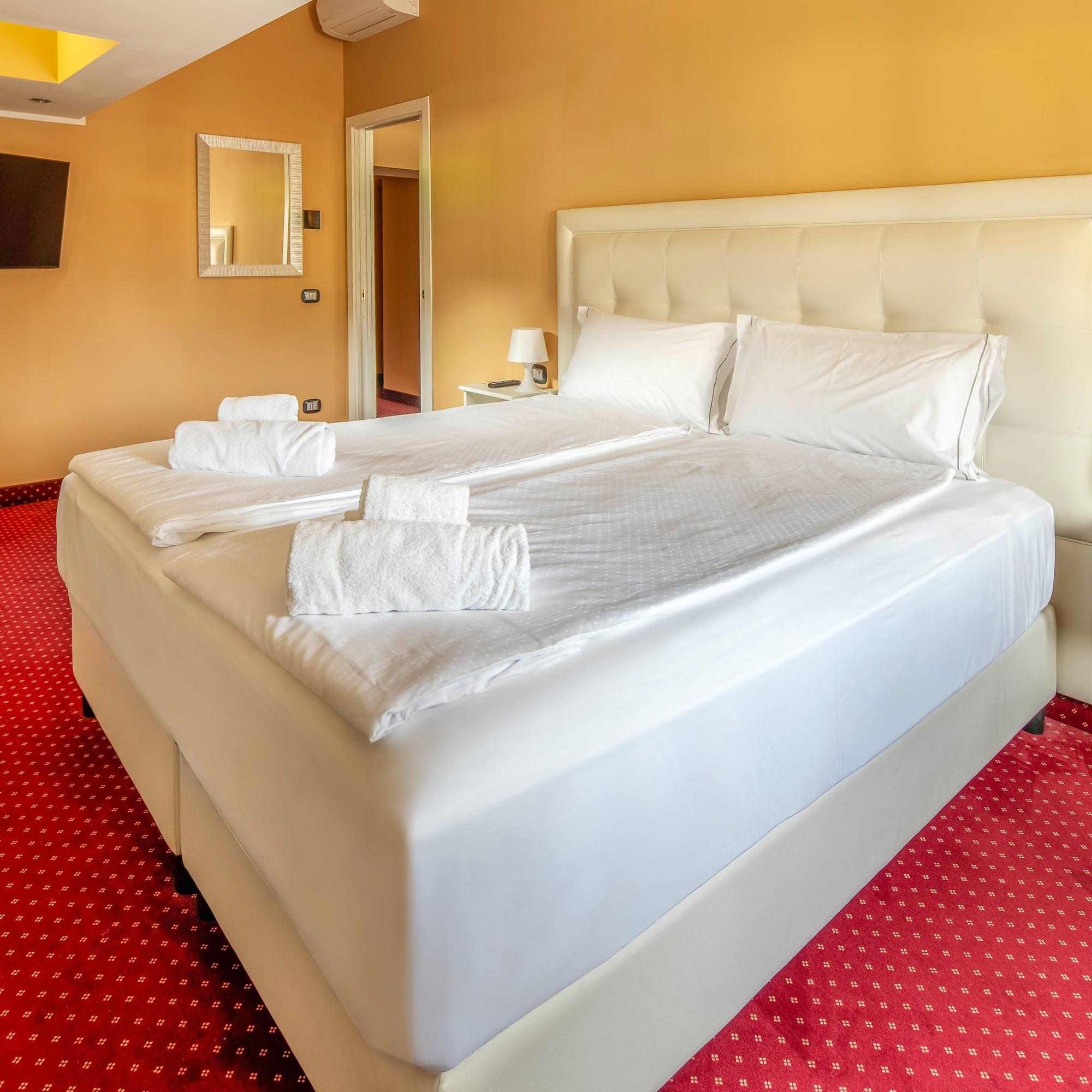 Hotel room with queen bed, white linen, red carpet, and orange walls.