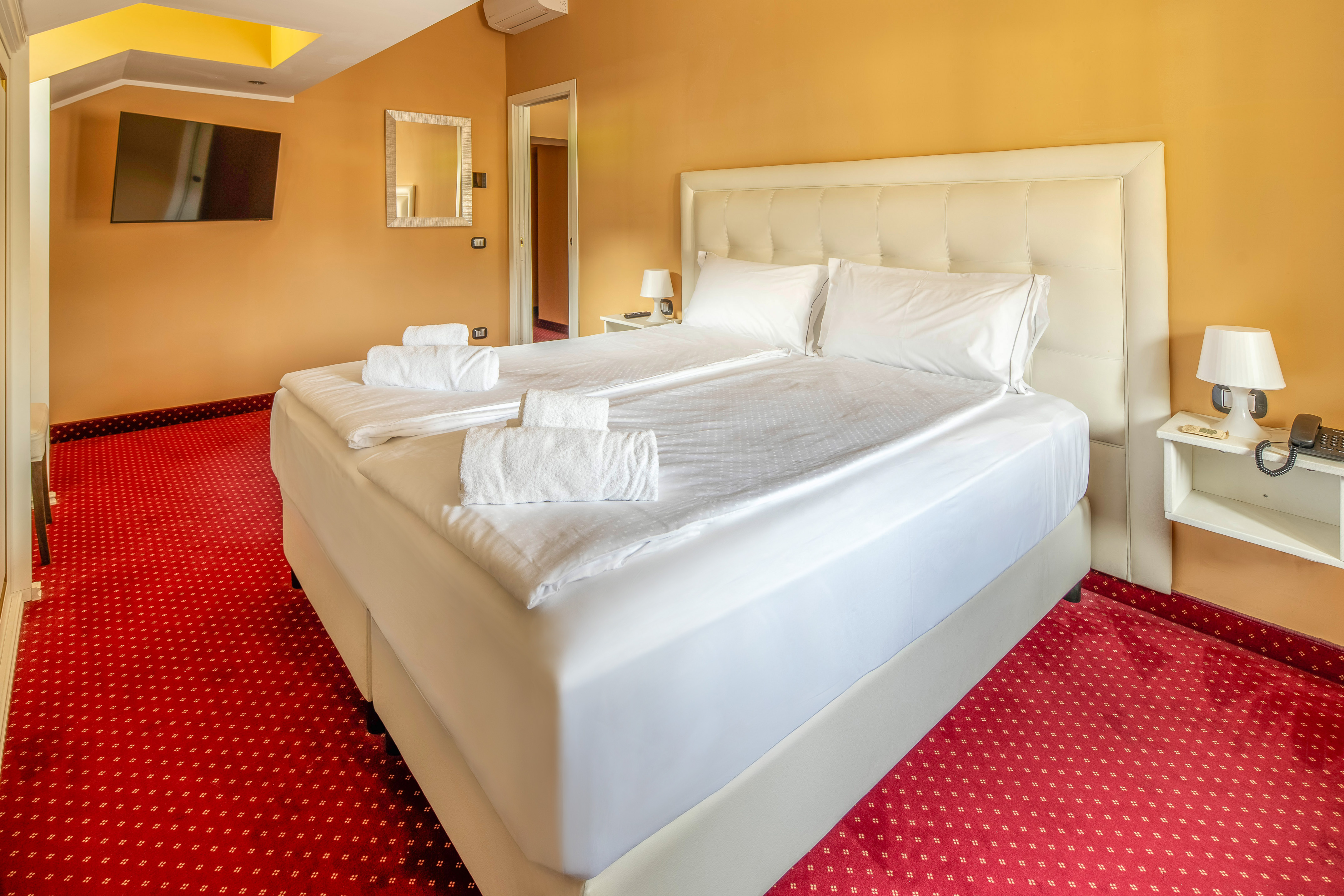Hotel room with queen bed, white linen, red carpet, and orange walls.
