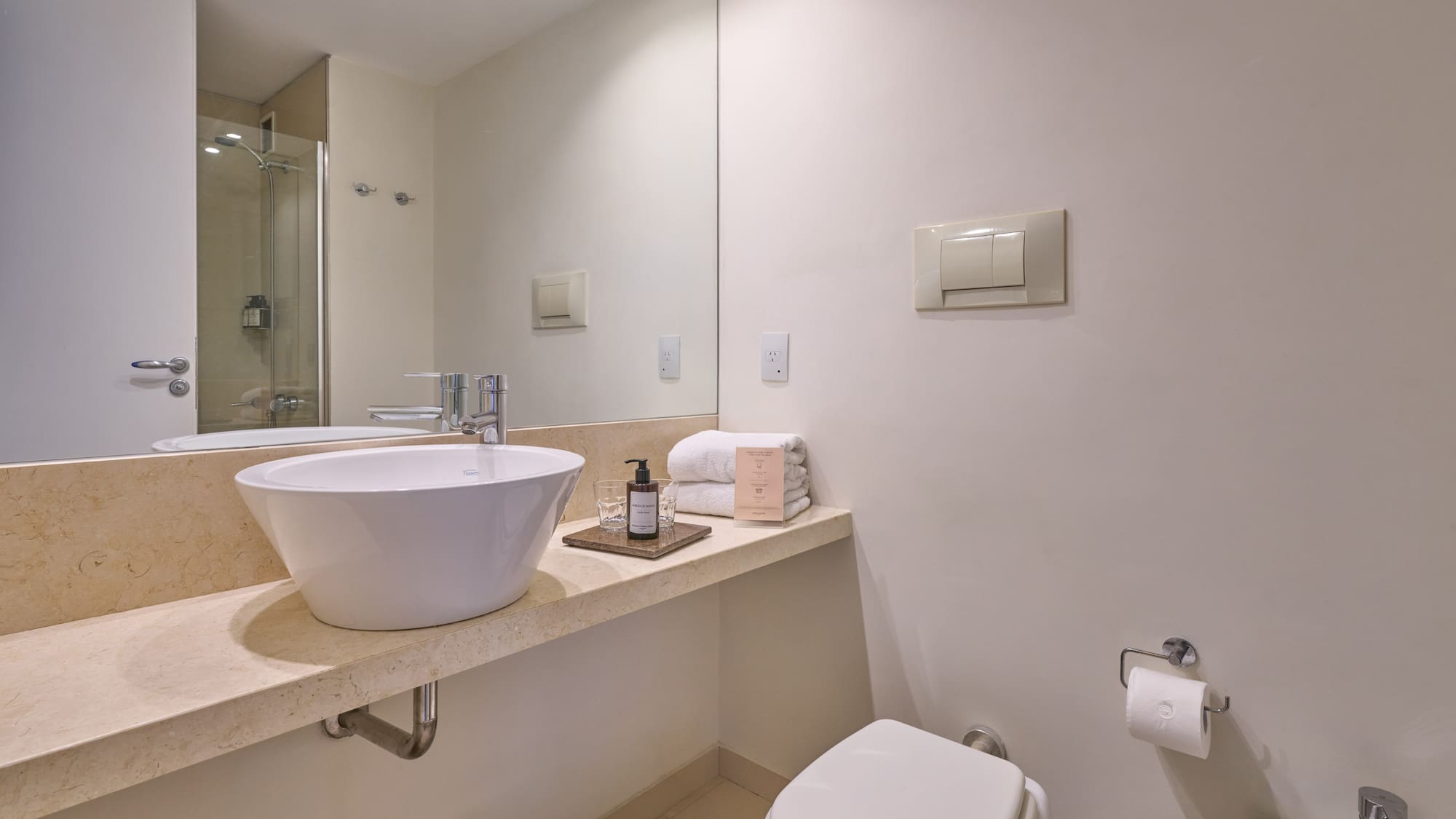 Modern bathroom with white vessel sink, marble vanity, toilet, and shower reflection.