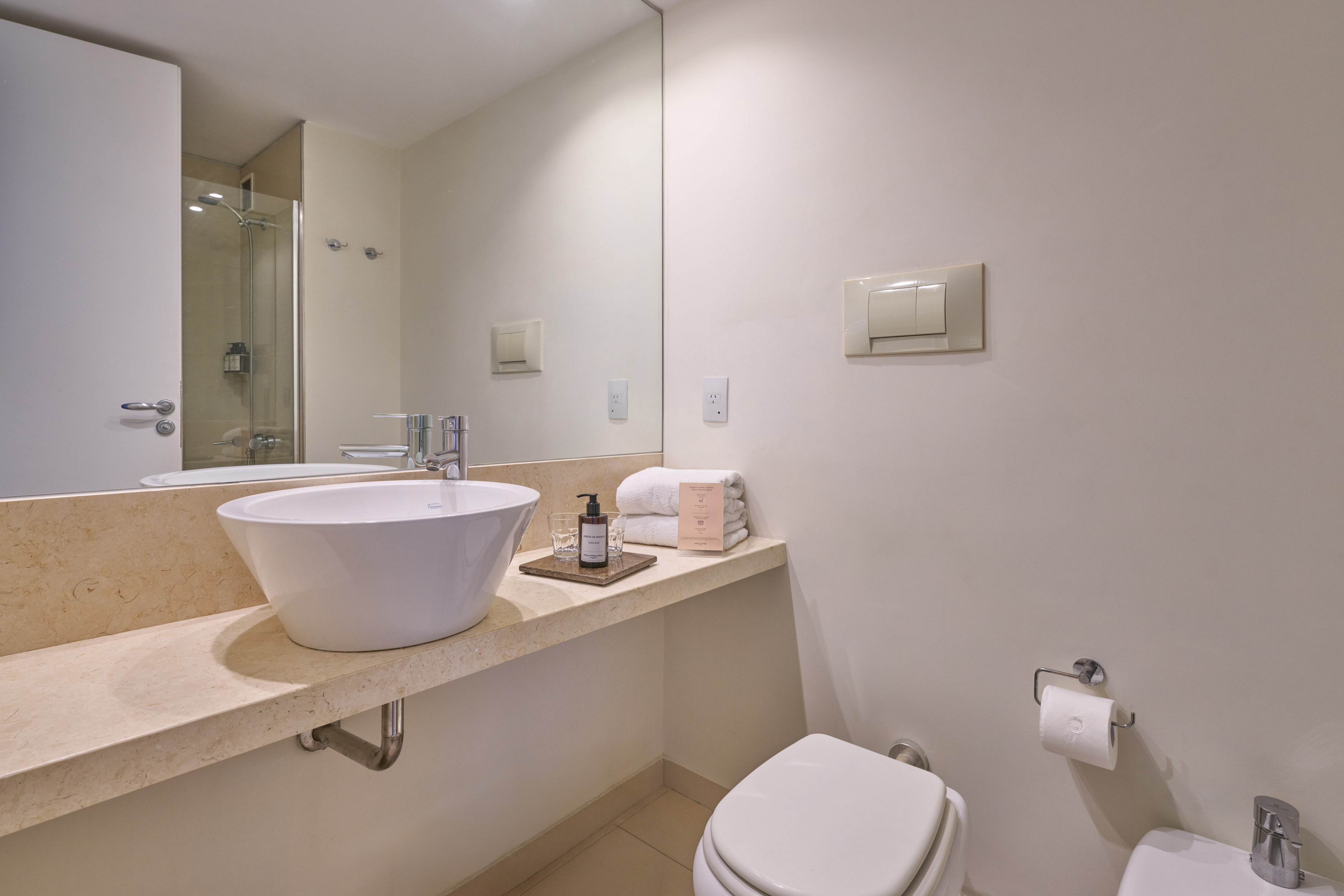 Modern bathroom with white vessel sink, marble vanity, toilet, and shower reflection.