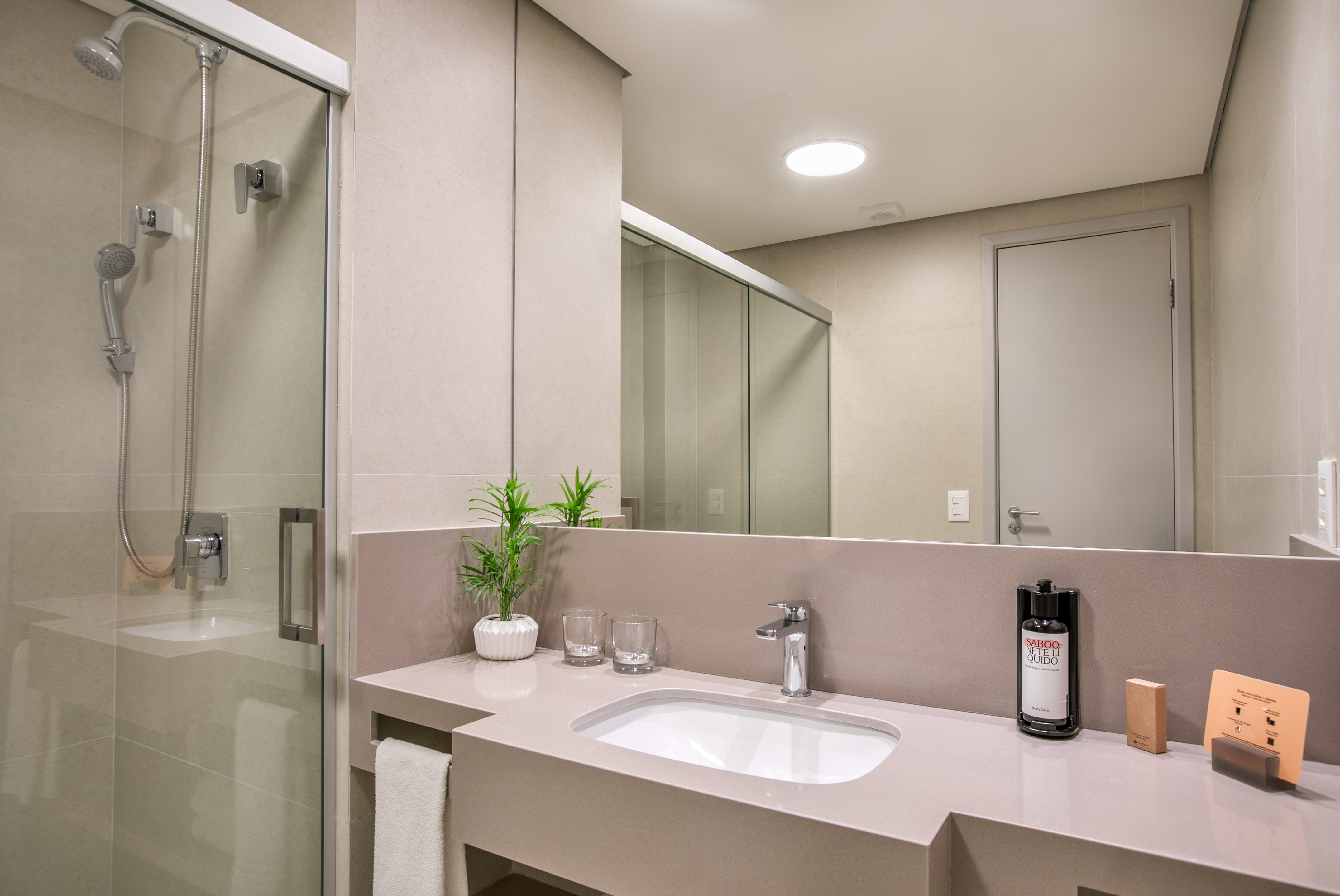 Modern hotel bathroom with glass shower, vanity sink, mirror, and beige tiles.