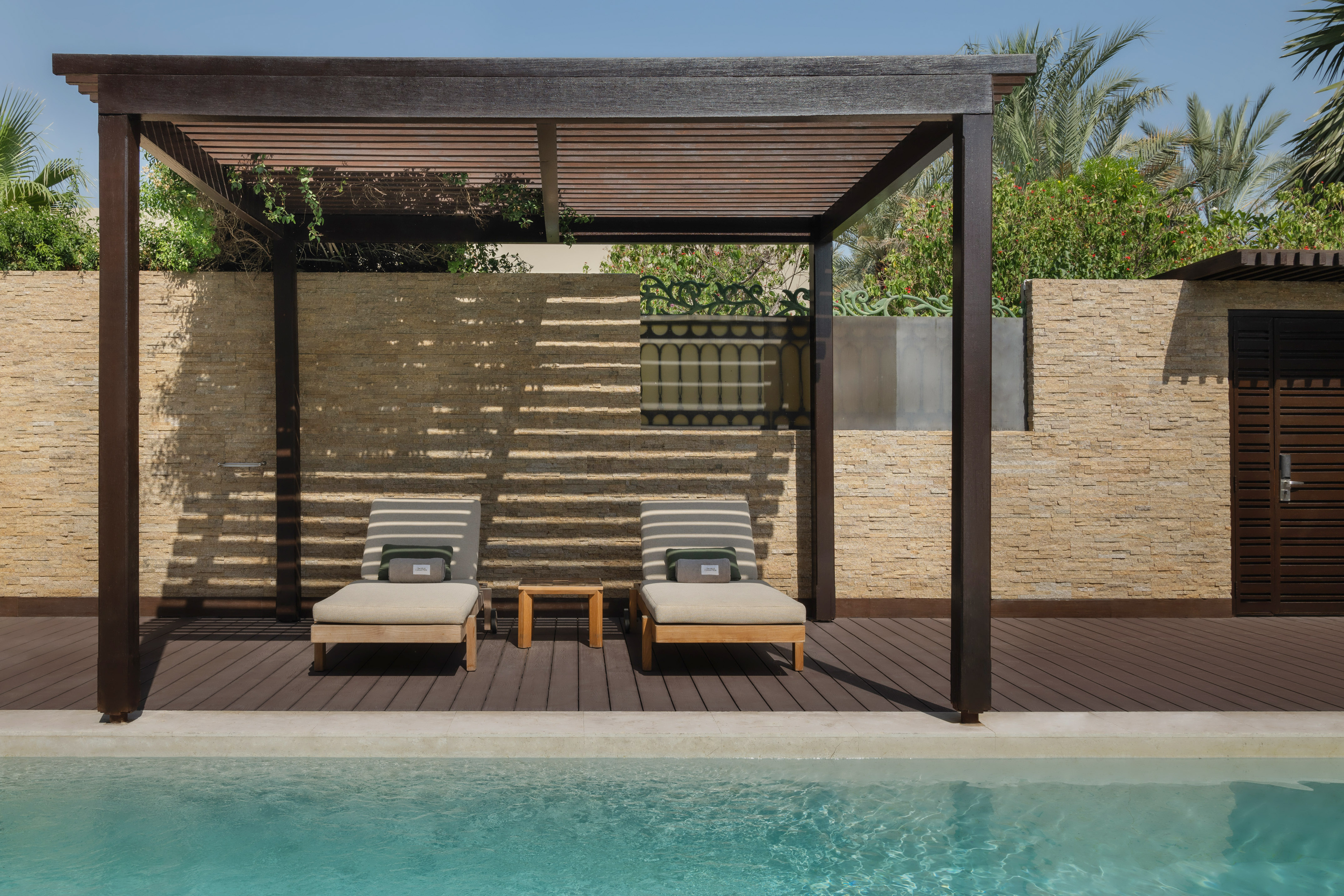 Poolside deck with two lounge chairs under a wooden pergola, by a clear pool.