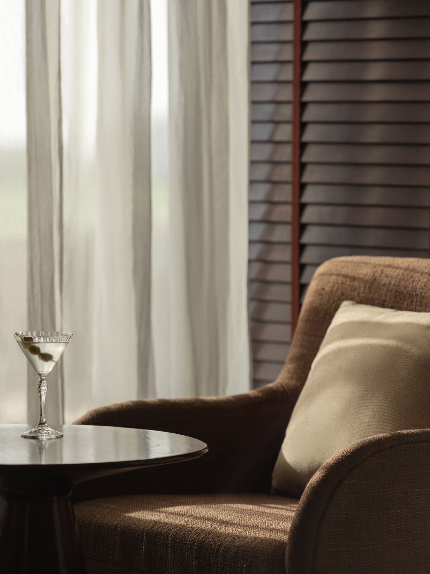Martini cocktail on side table next to armchair by window with sheer curtains.