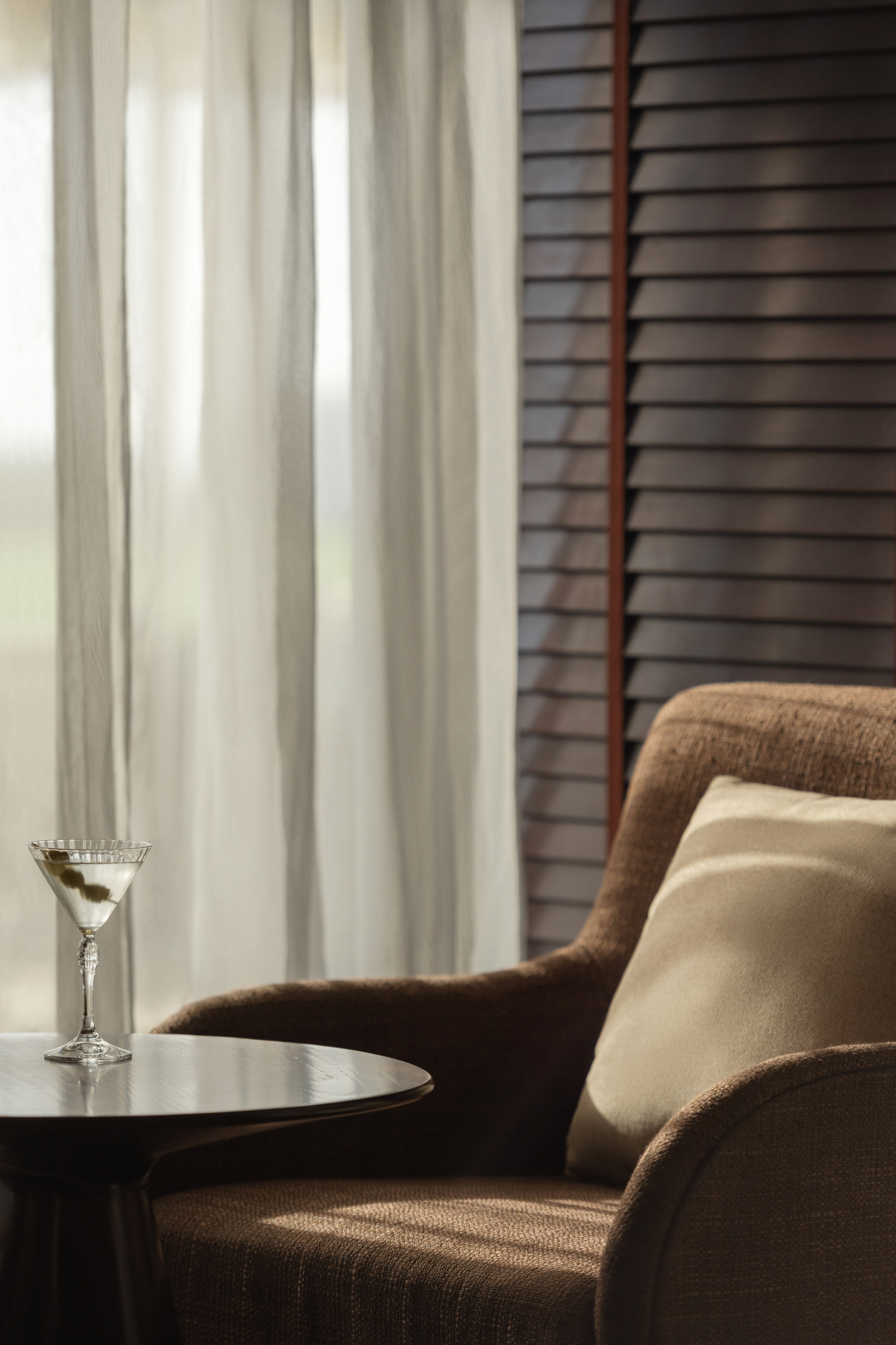 Martini cocktail on side table next to armchair by window with sheer curtains.
