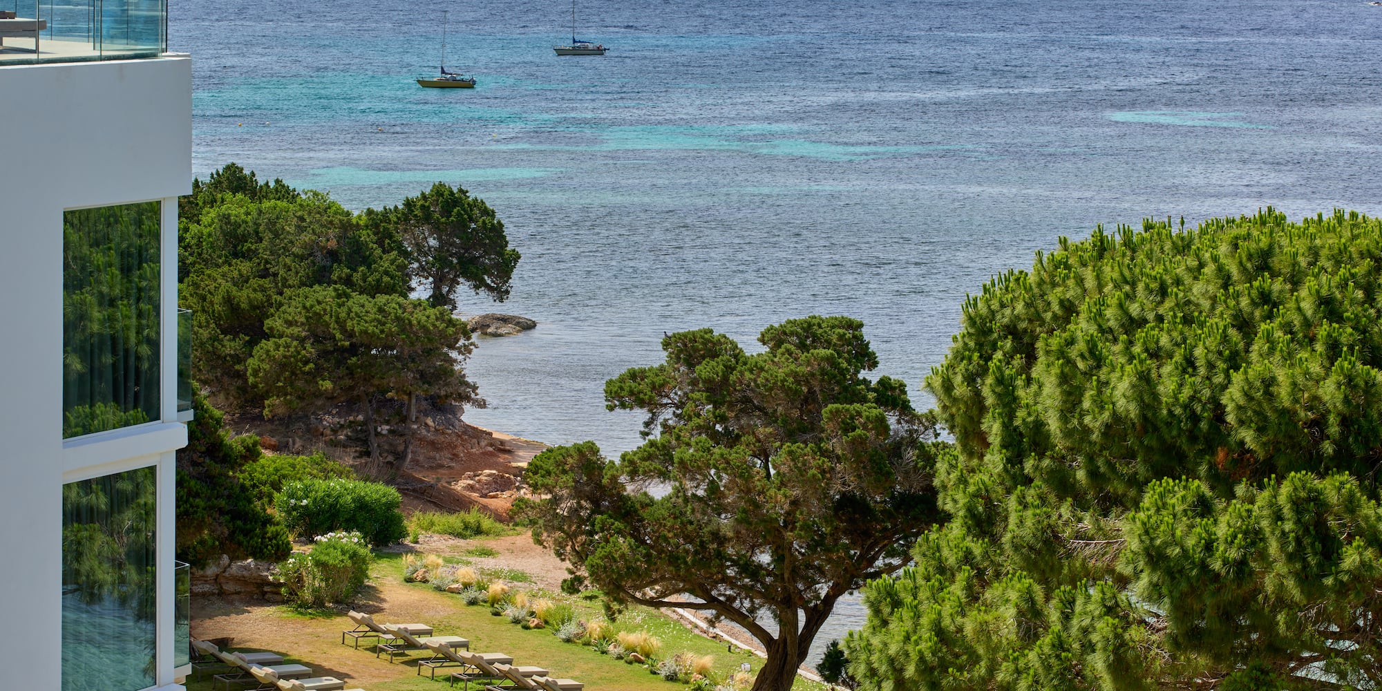 Modern resort with sea view, sun loungers, green trees, and two sailboats.