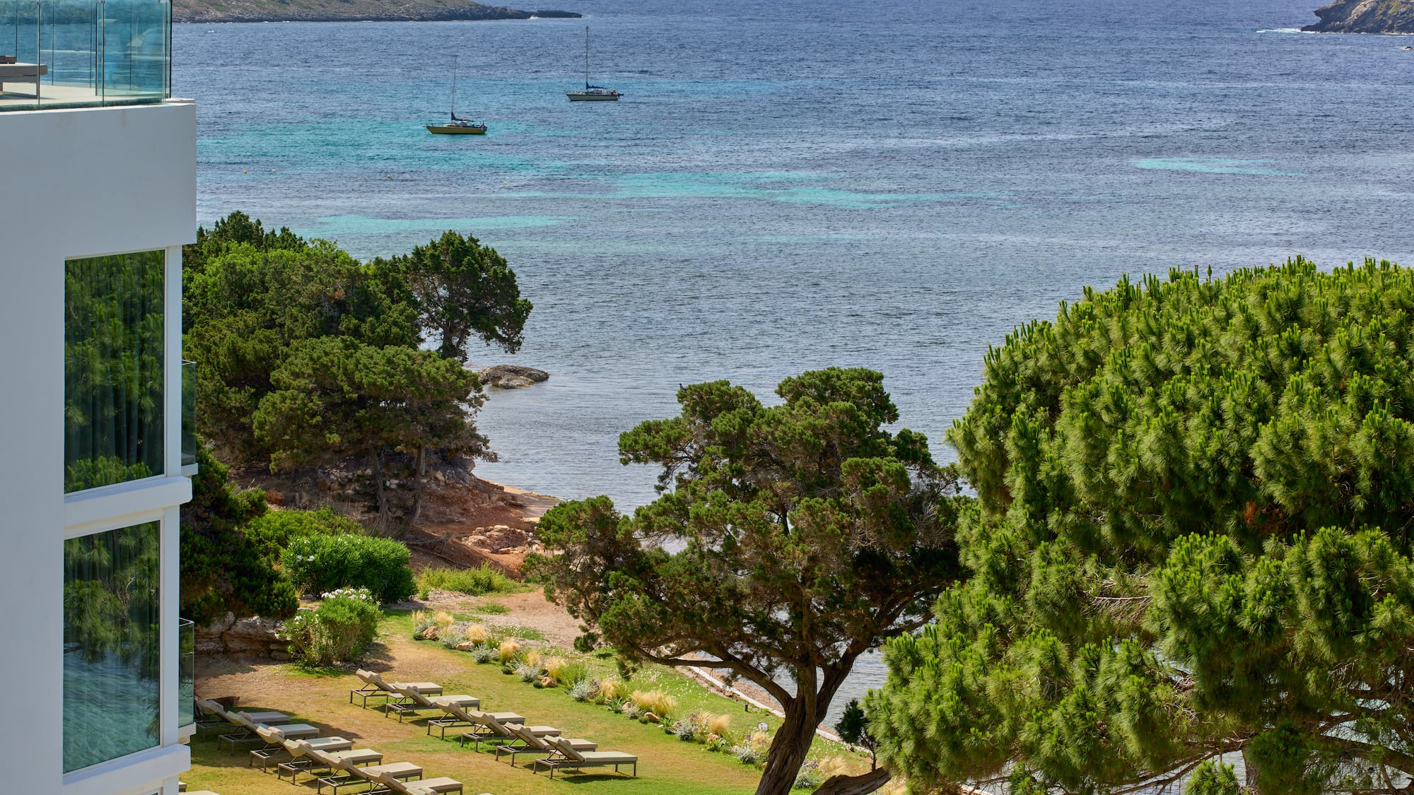 Modern resort with sea view, sun loungers, green trees, and two sailboats.
