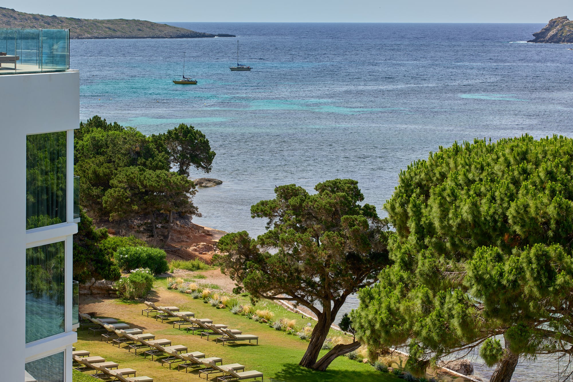 Modern resort with sea view, sun loungers, green trees, and two sailboats.