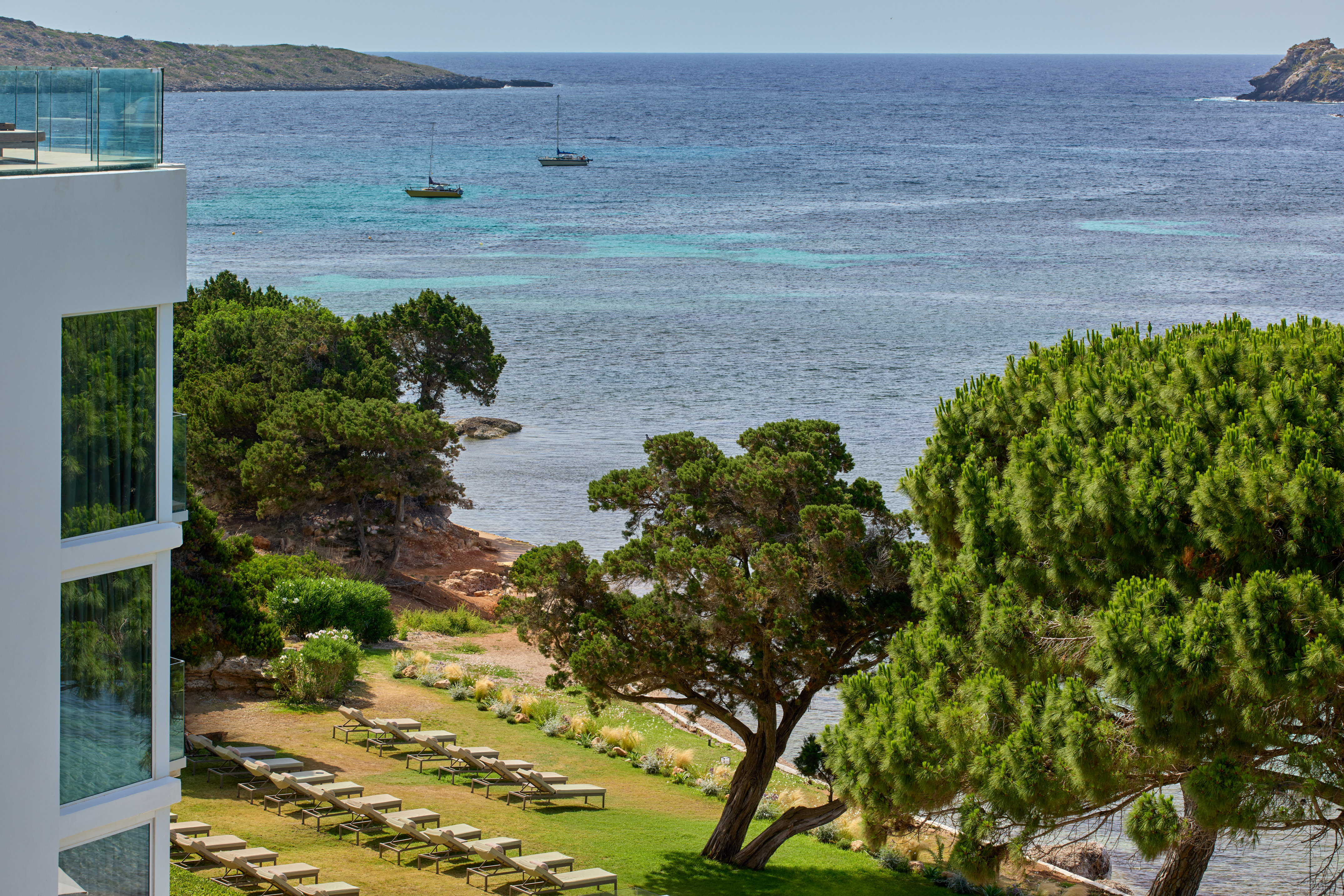 Modern resort with sea view, sun loungers, green trees, and two sailboats.