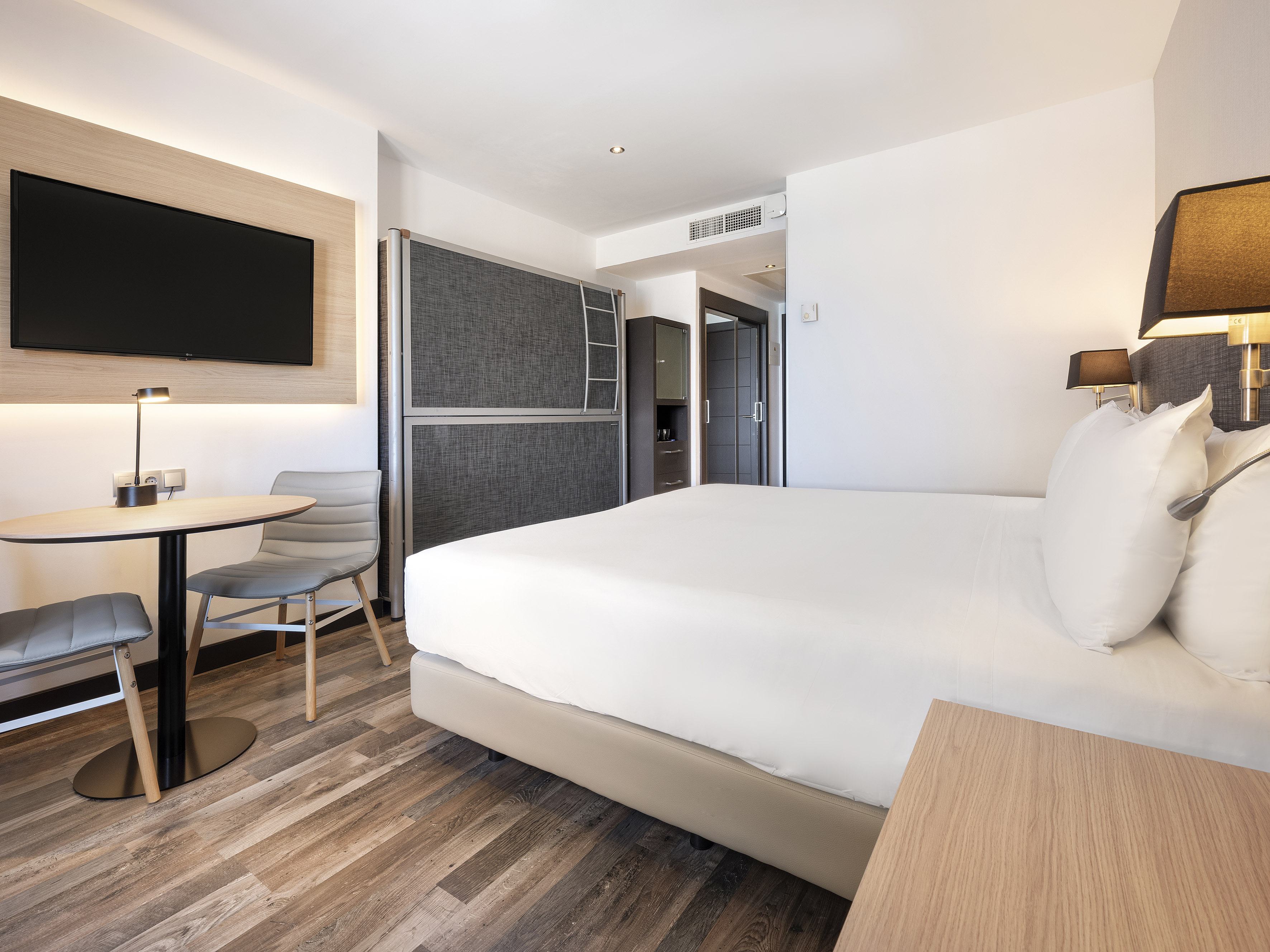 Modern hotel room with queen bed, wooden floor, wall-mounted TV, and seating area.