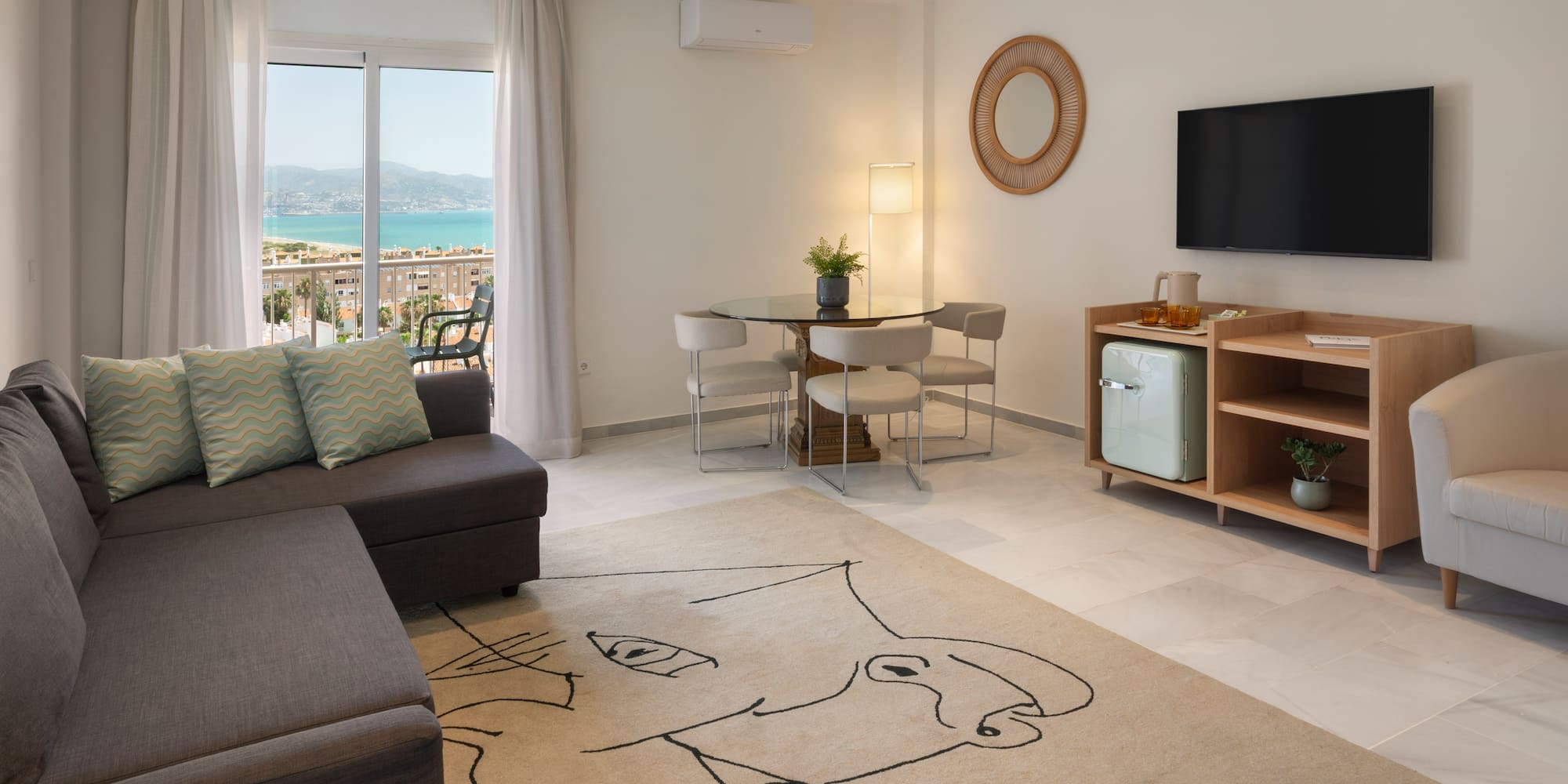 Salón moderno con sofá, mesa de comedor, TV y balcón con vistas al mar.