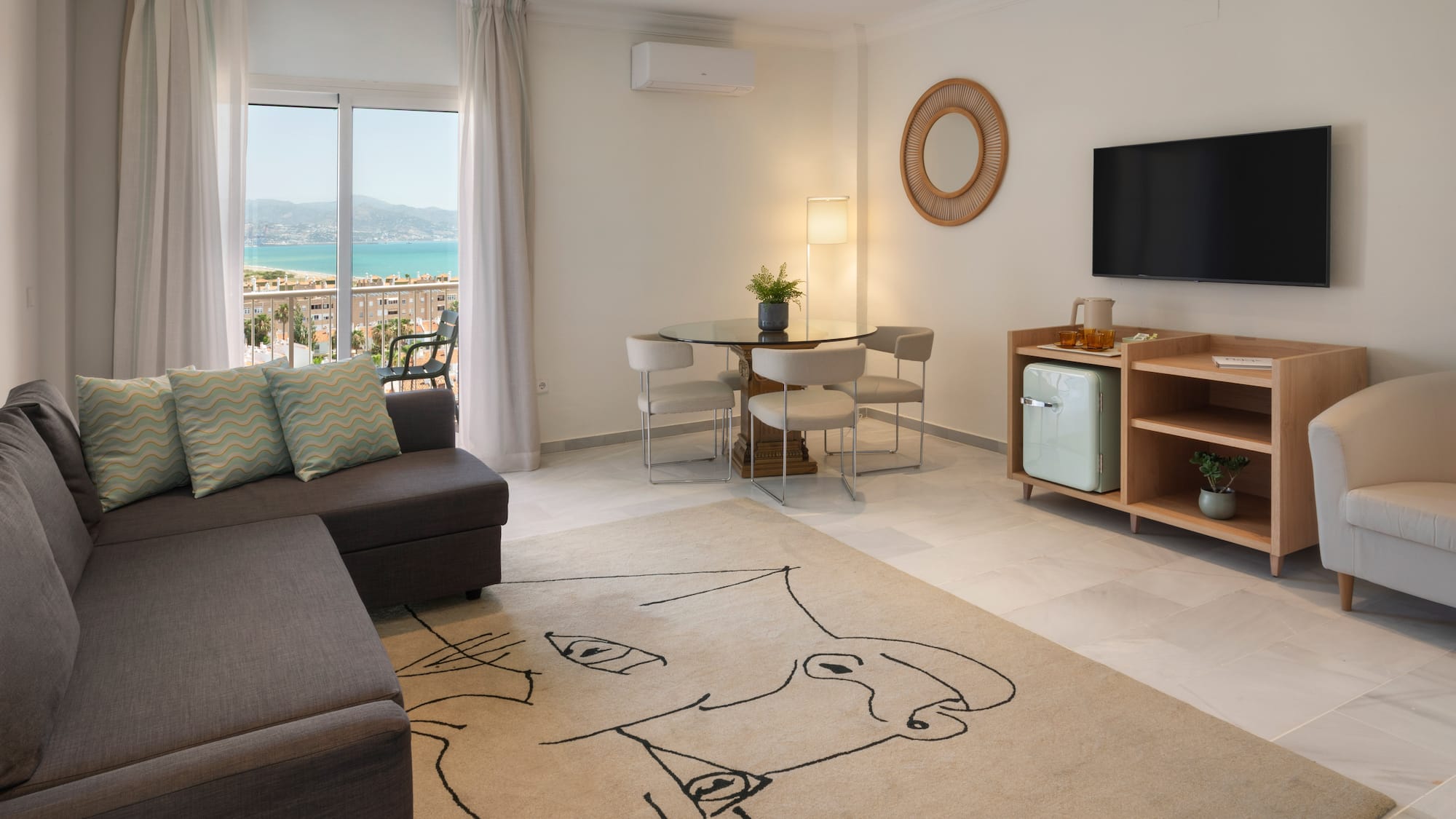 Salón moderno con sofá, mesa de comedor, TV y balcón con vistas al mar.