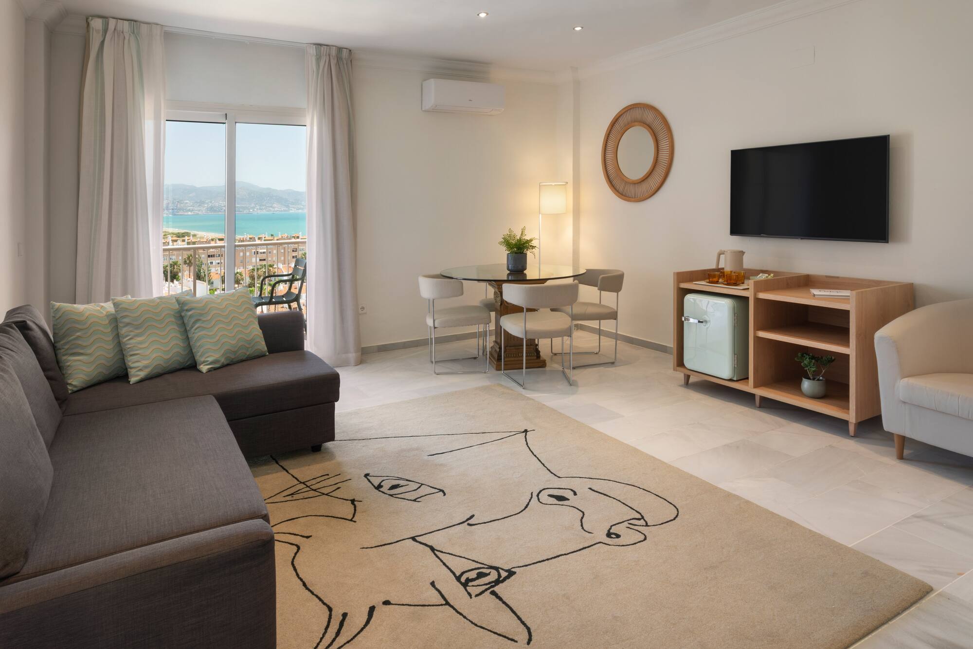 Salón moderno con sofá, mesa de comedor, TV y balcón con vistas al mar.