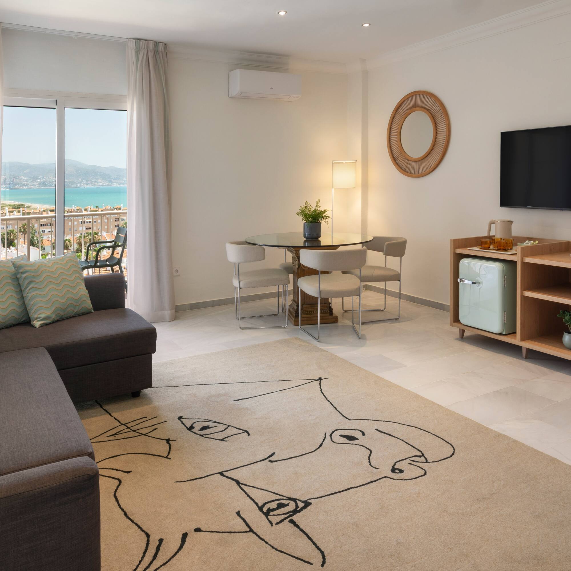 Salón moderno con sofá, mesa de comedor, TV y balcón con vistas al mar.