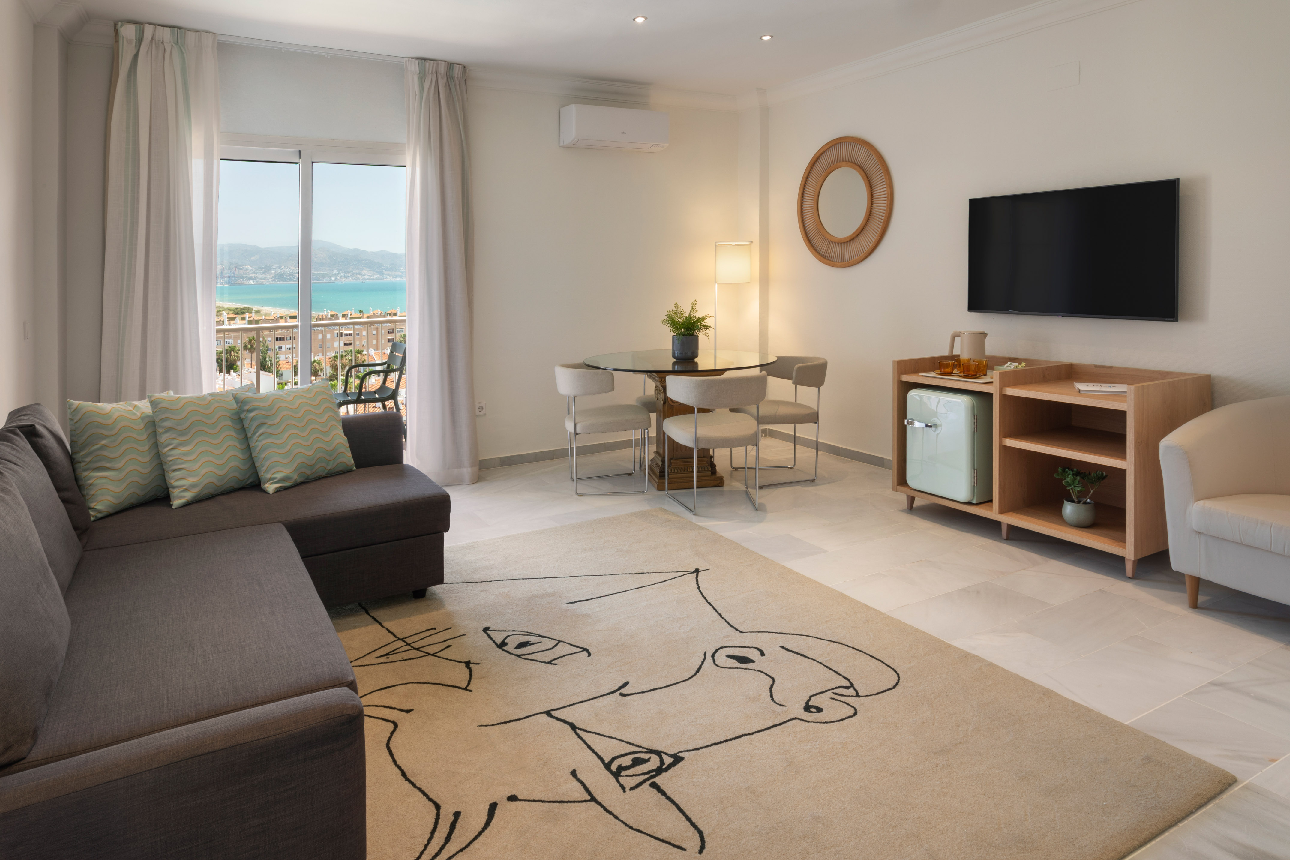 Salón moderno con sofá, mesa de comedor, TV y balcón con vistas al mar.