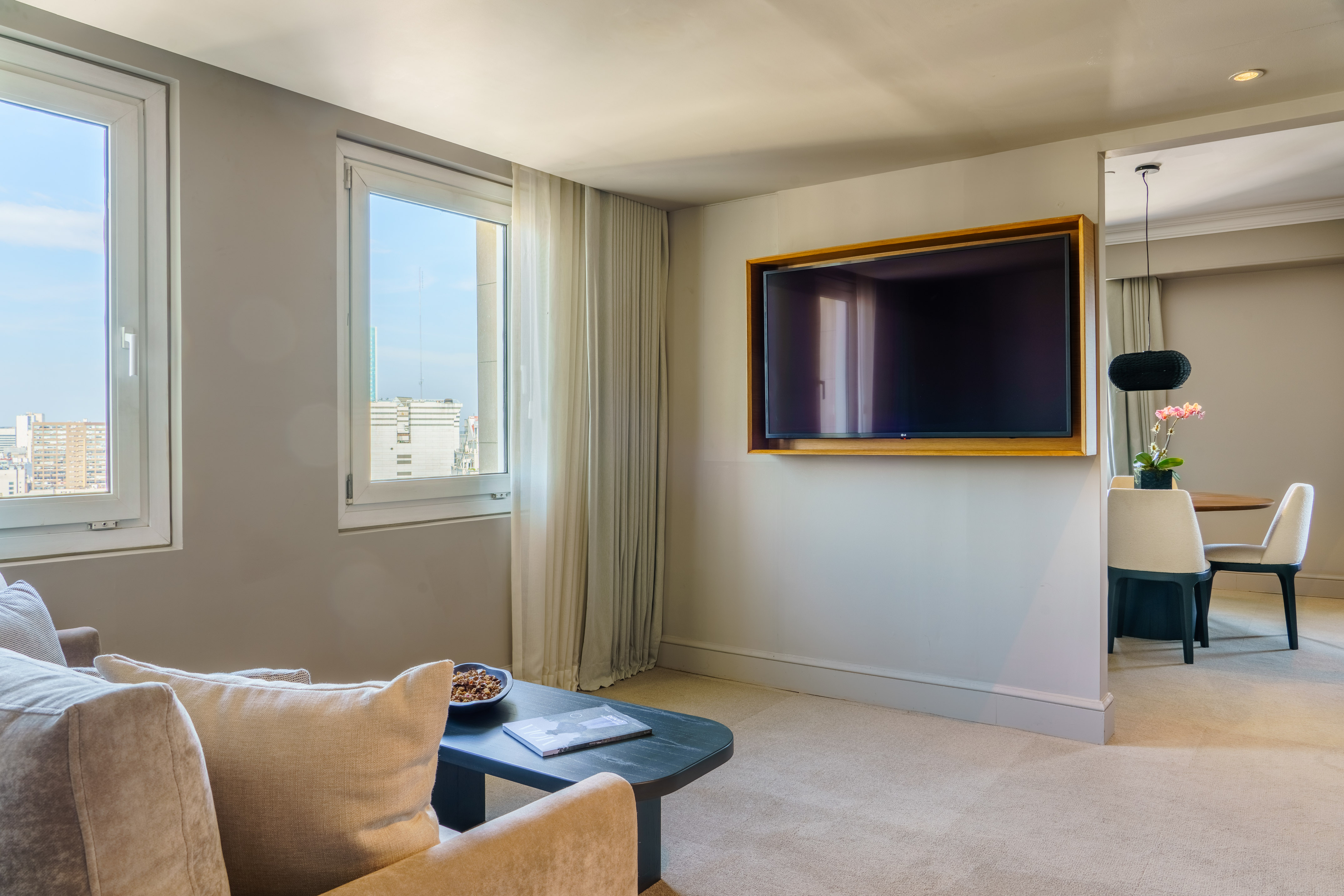 Hotel suite living room with flat-screen TV, sofa, city view, and dining area.