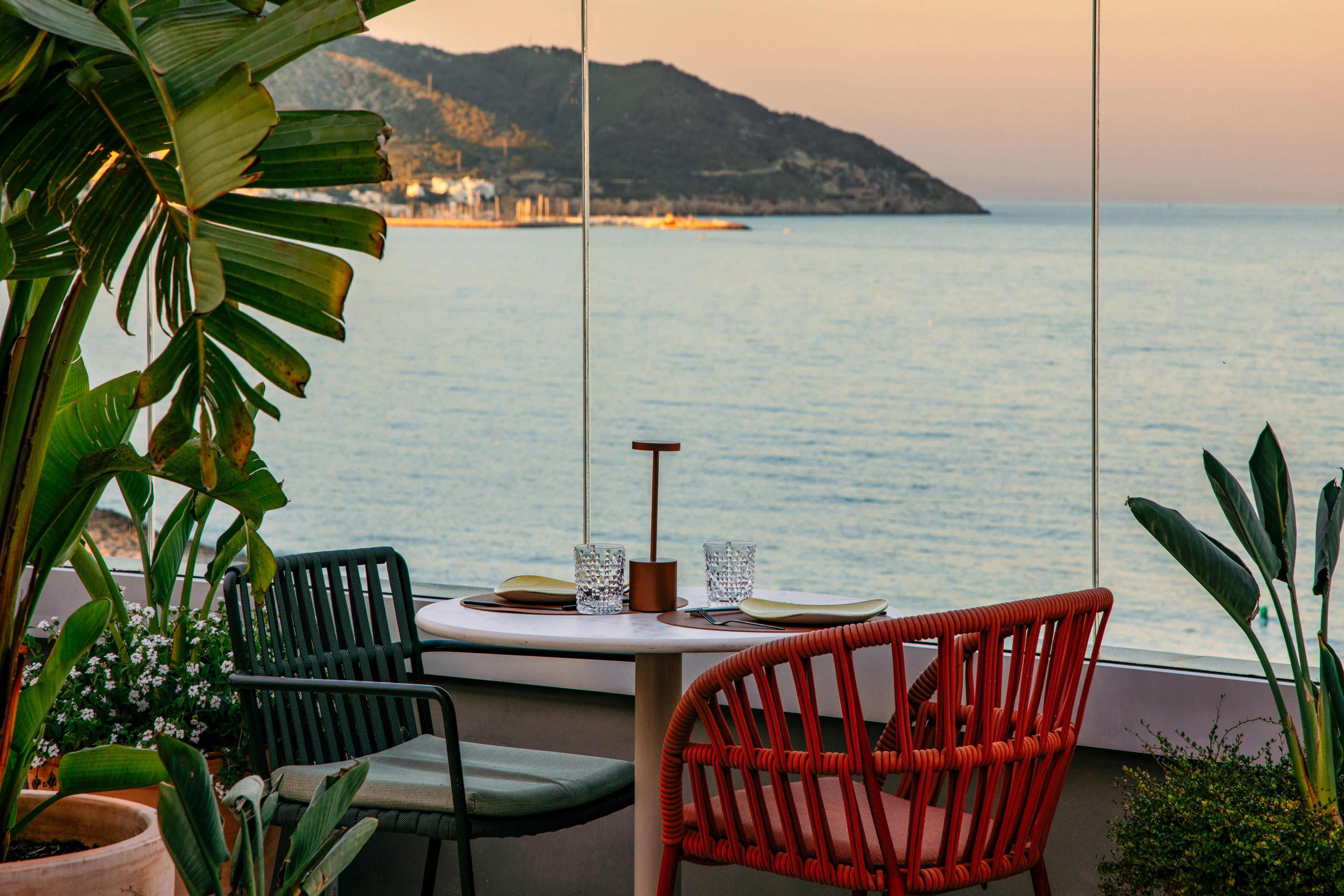 Terrace dining table overlooking the sea at sunset, with tropical plants.