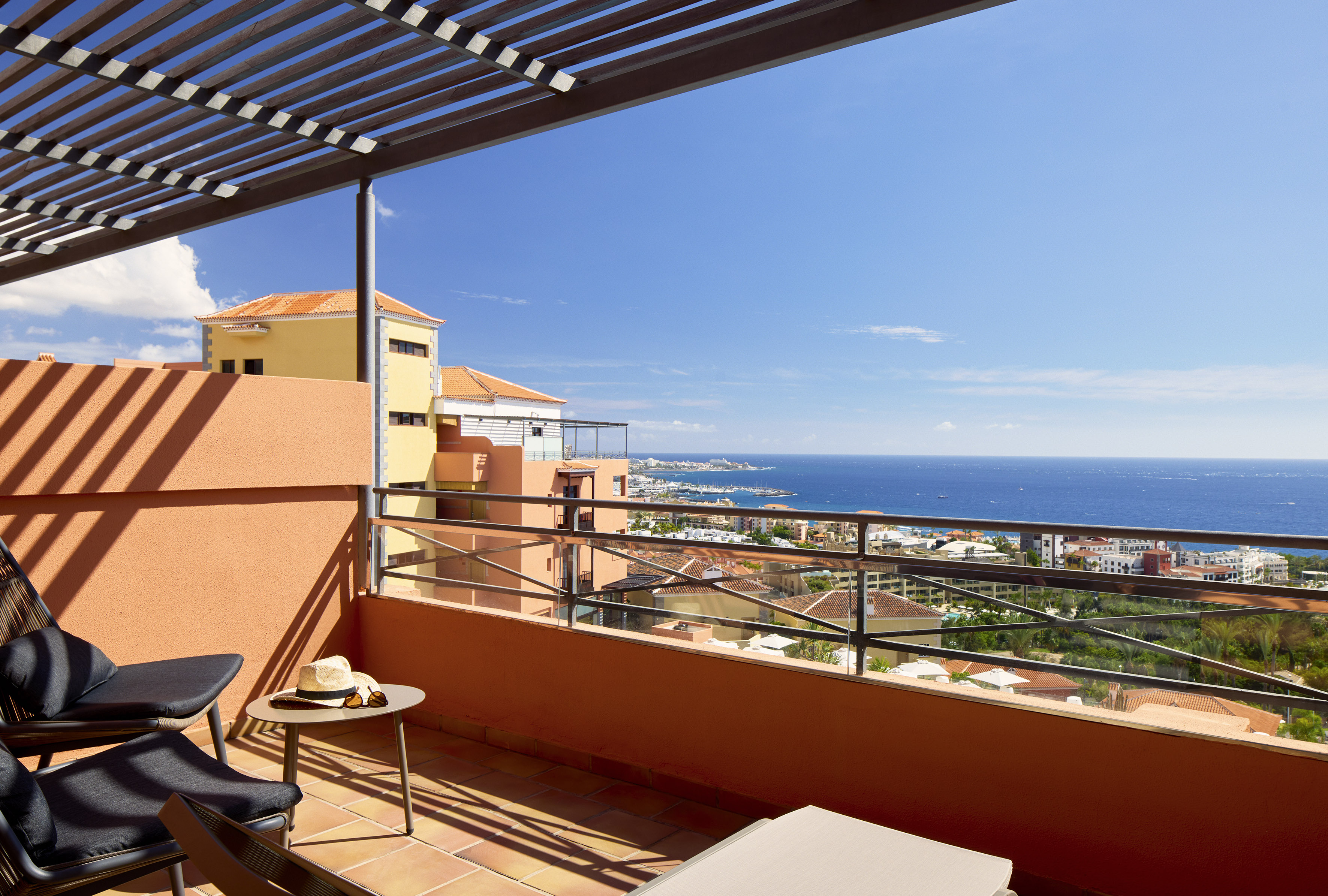 Sunny balcony with chairs, pergola, and panoramic sea view of a coastal resort.