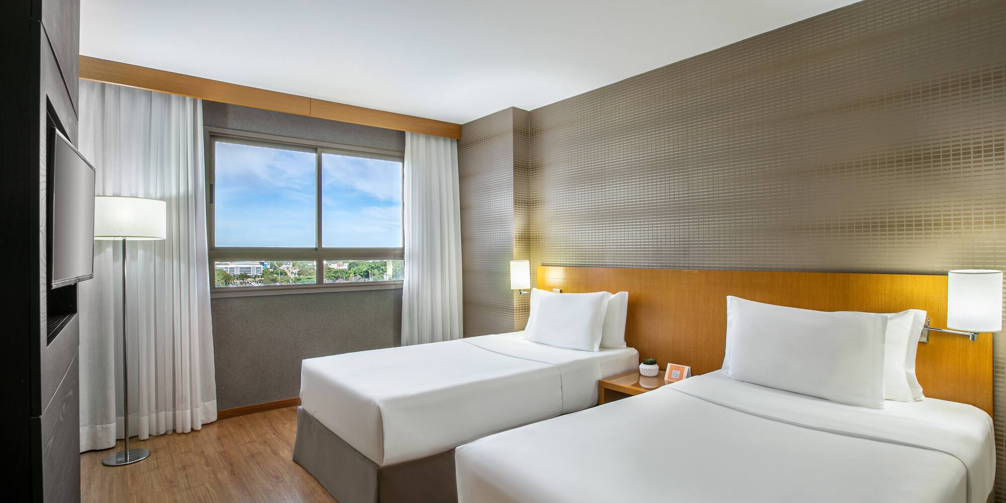 Modern hotel room with two twin beds, wooden floor, window, and TV.
