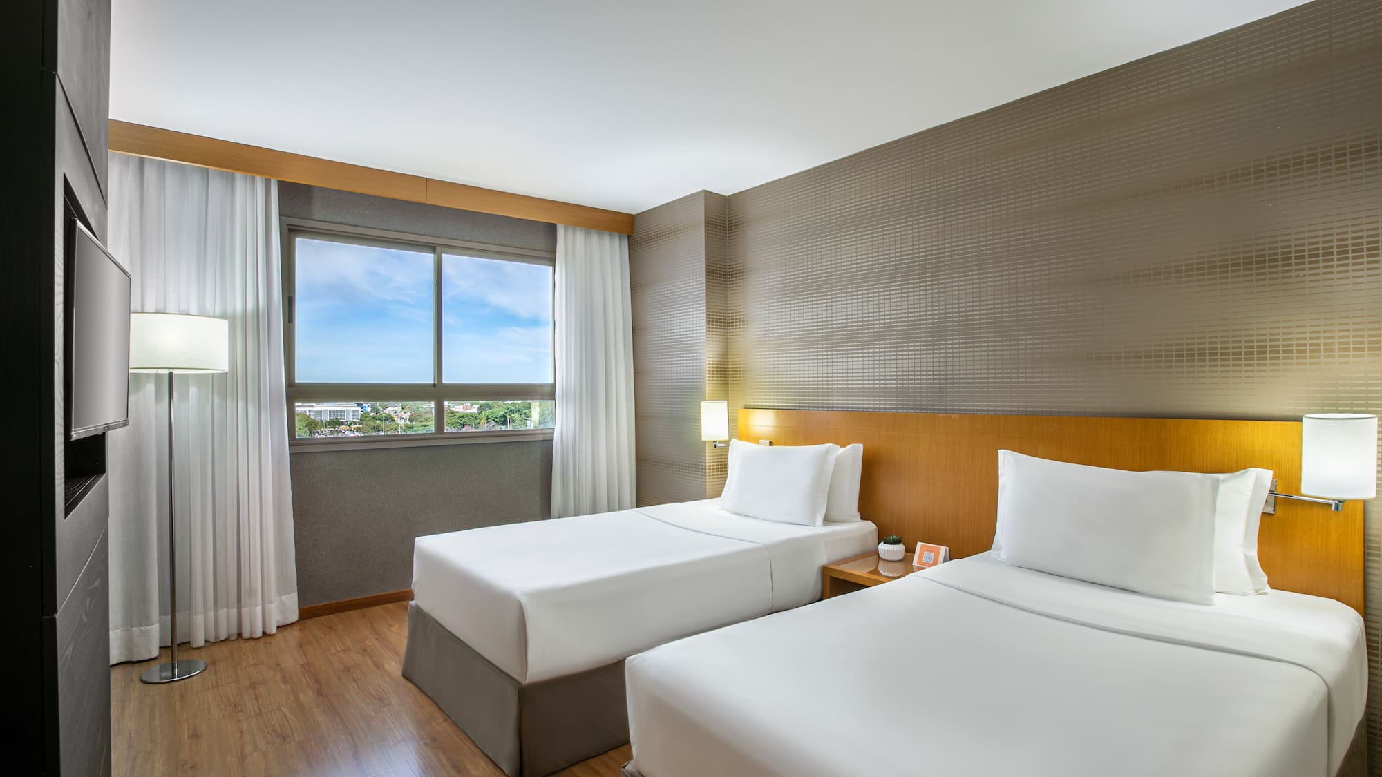 Modern hotel room with two twin beds, wooden floor, window, and TV.