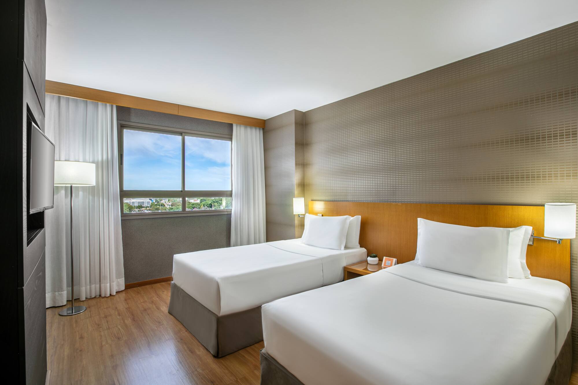 Modern hotel room with two twin beds, wooden floor, window, and TV.