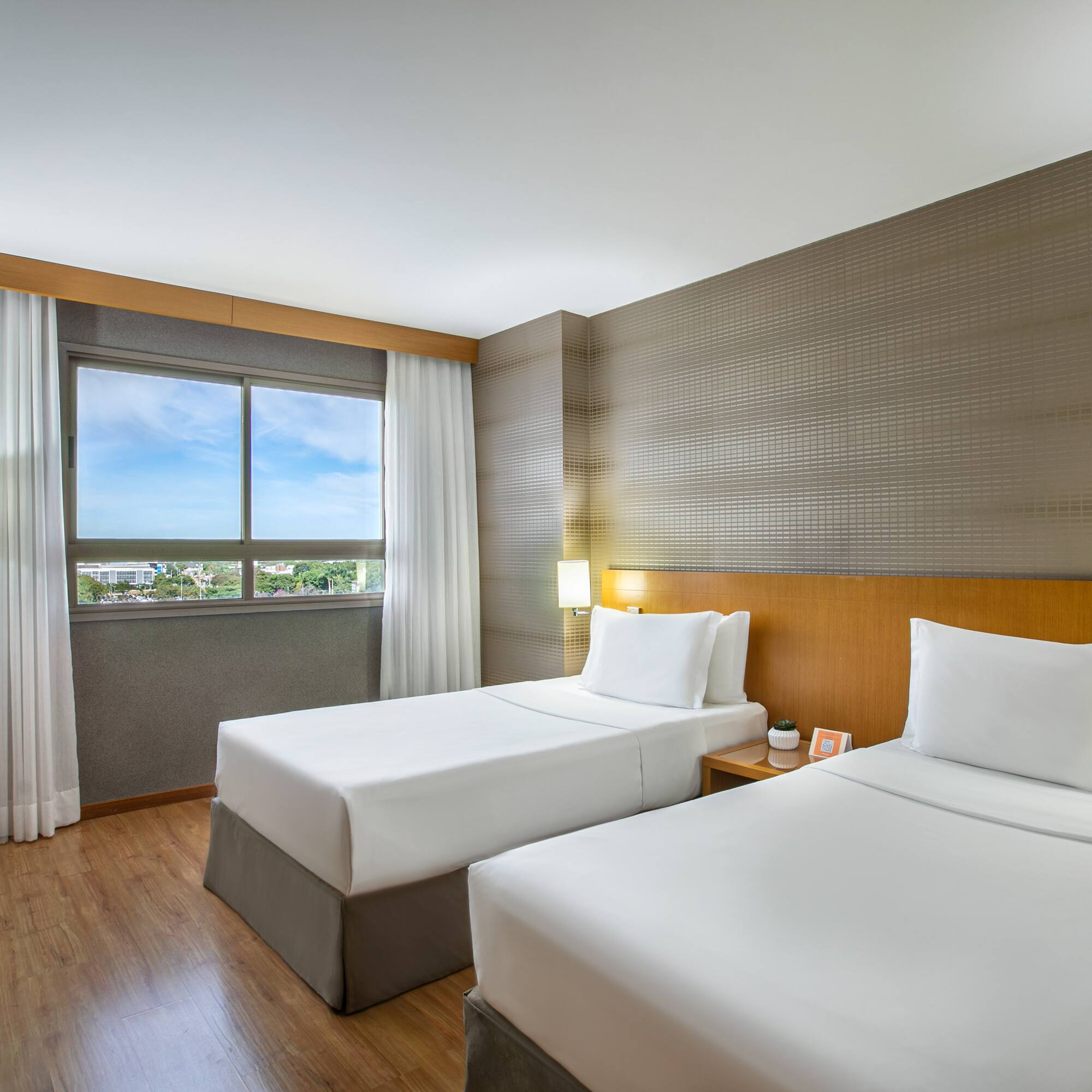 Modern hotel room with two twin beds, wooden floor, window, and TV.
