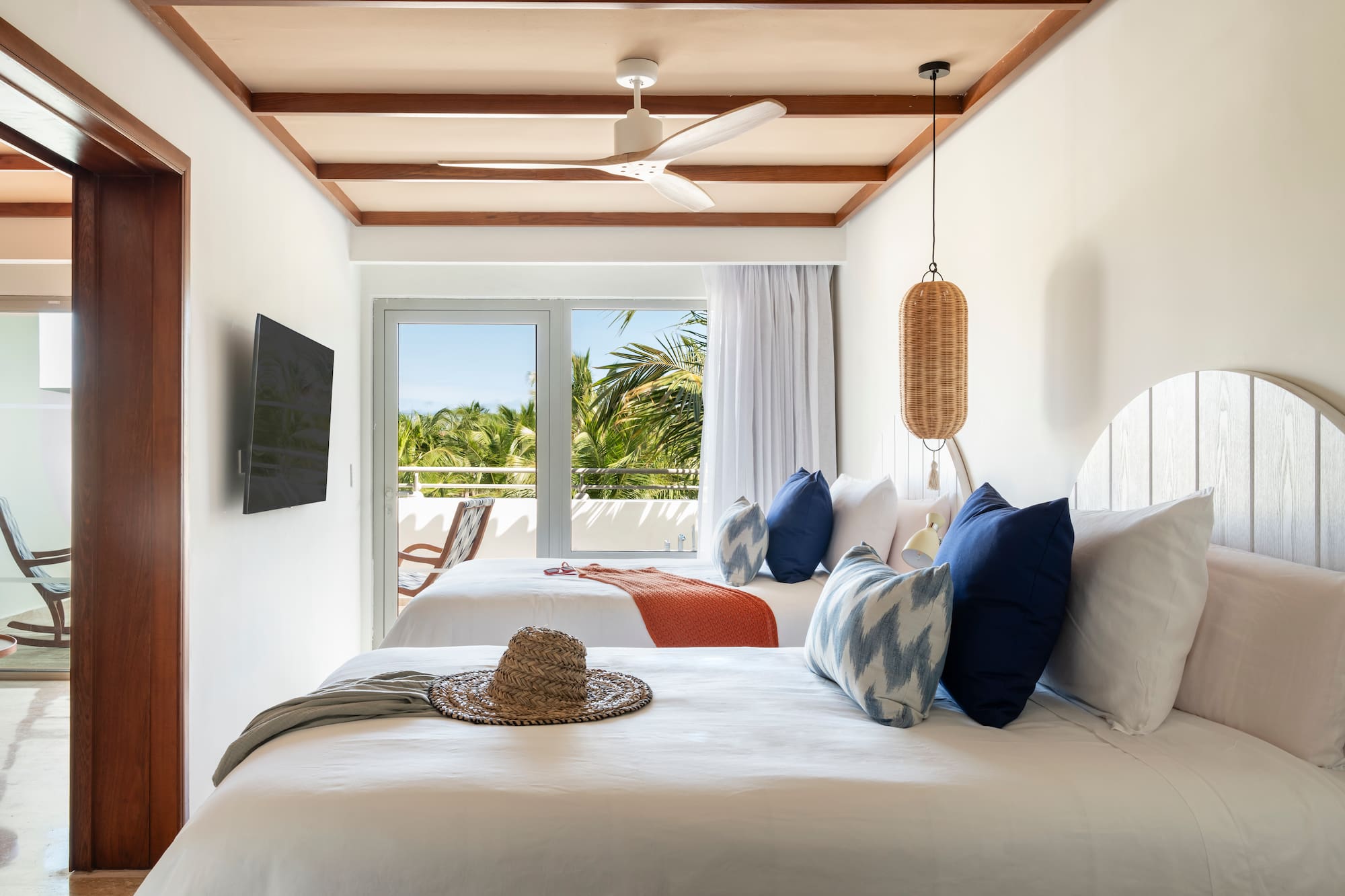 Tropical hotel room, two beds, balcony with palm tree view, ceiling fan.