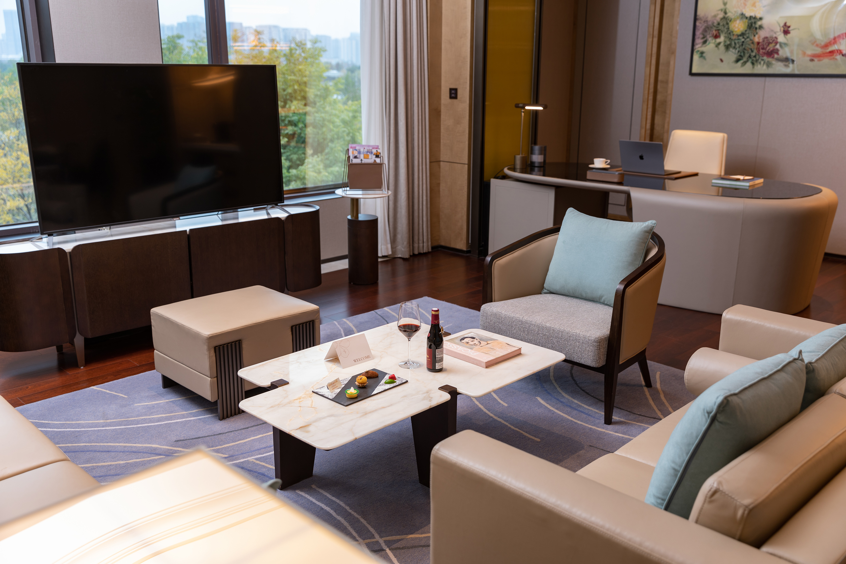 Modern hotel suite living room with sofa, coffee table, large TV, and desk setup.