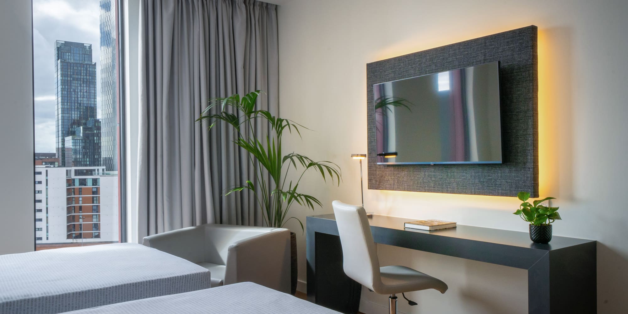 Modern hotel room with twin beds, city view, desk, and wall-mounted TV.