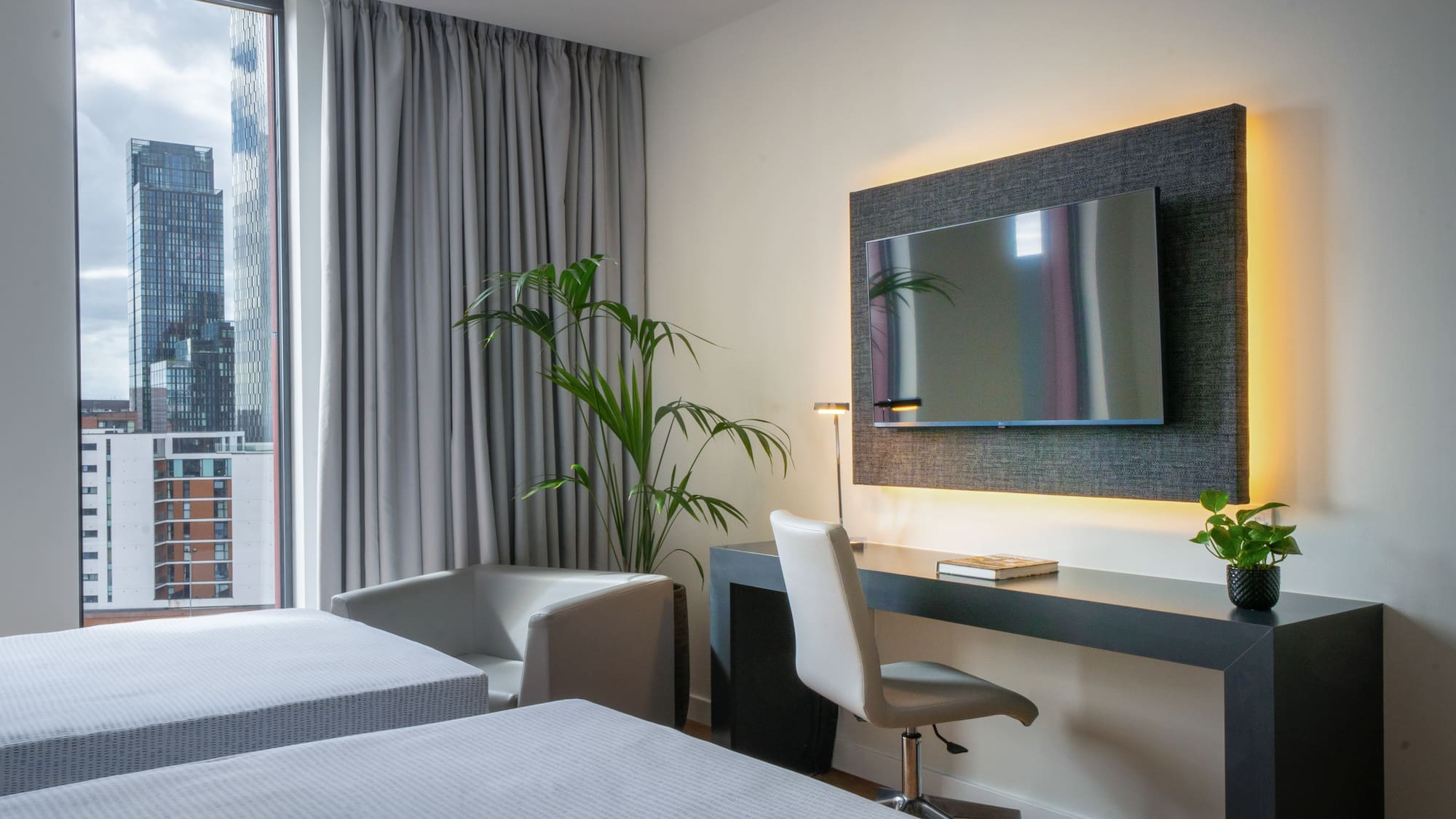 Modern hotel room with twin beds, city view, desk, and wall-mounted TV.