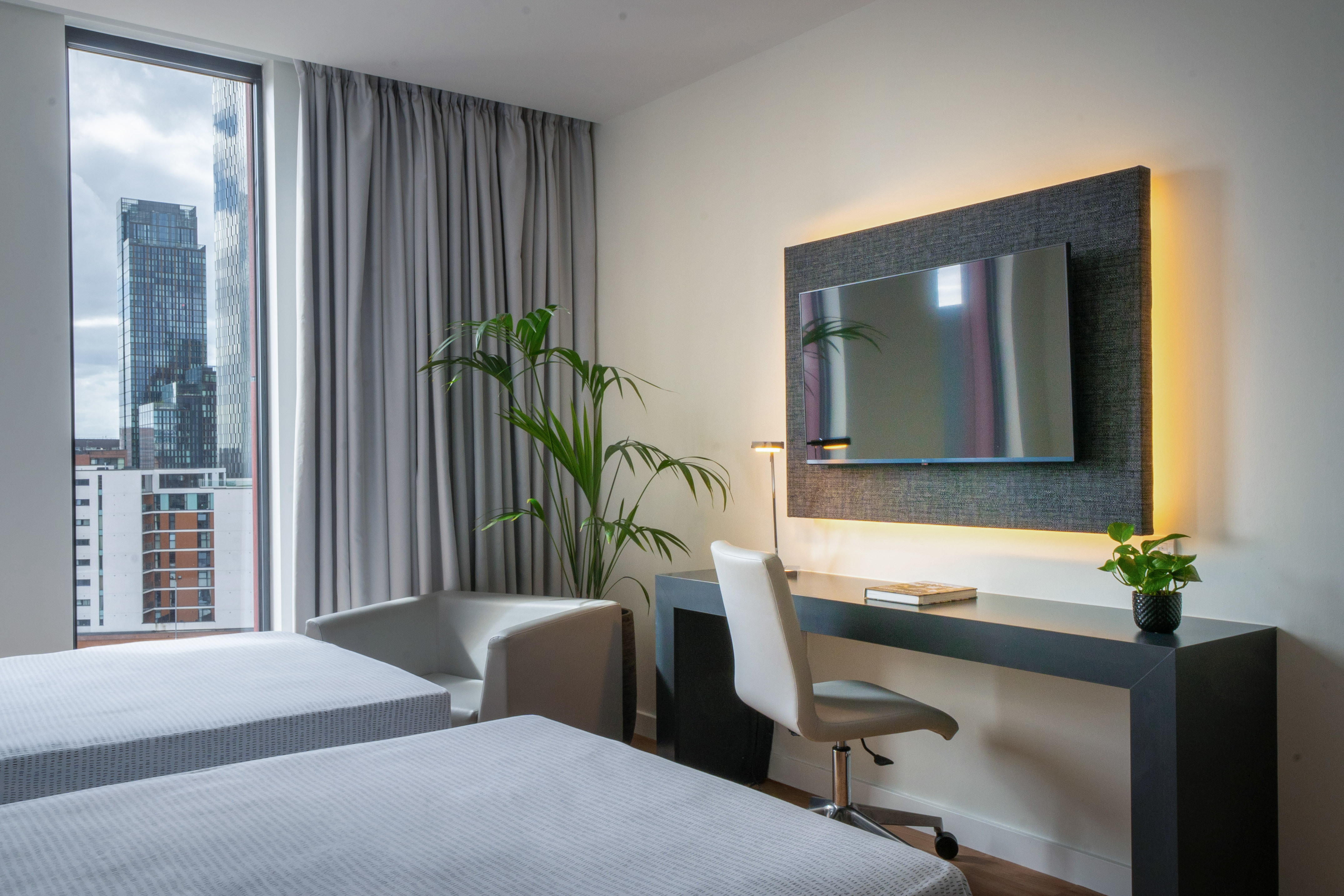 Modern hotel room with twin beds, city view, desk, and wall-mounted TV.