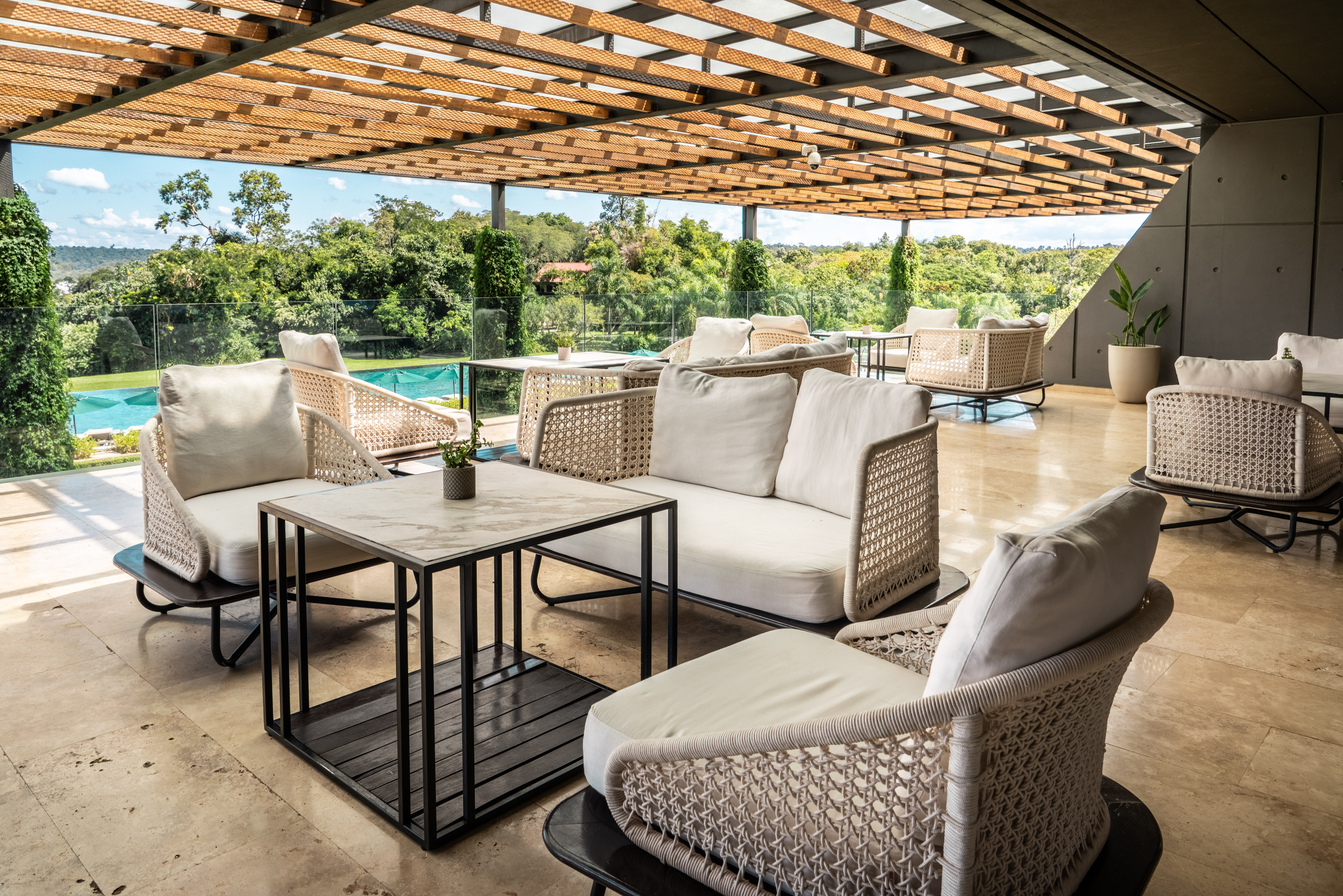 Outdoor lounge with modern rattan furniture, wooden pergola, pool, and tropical view.