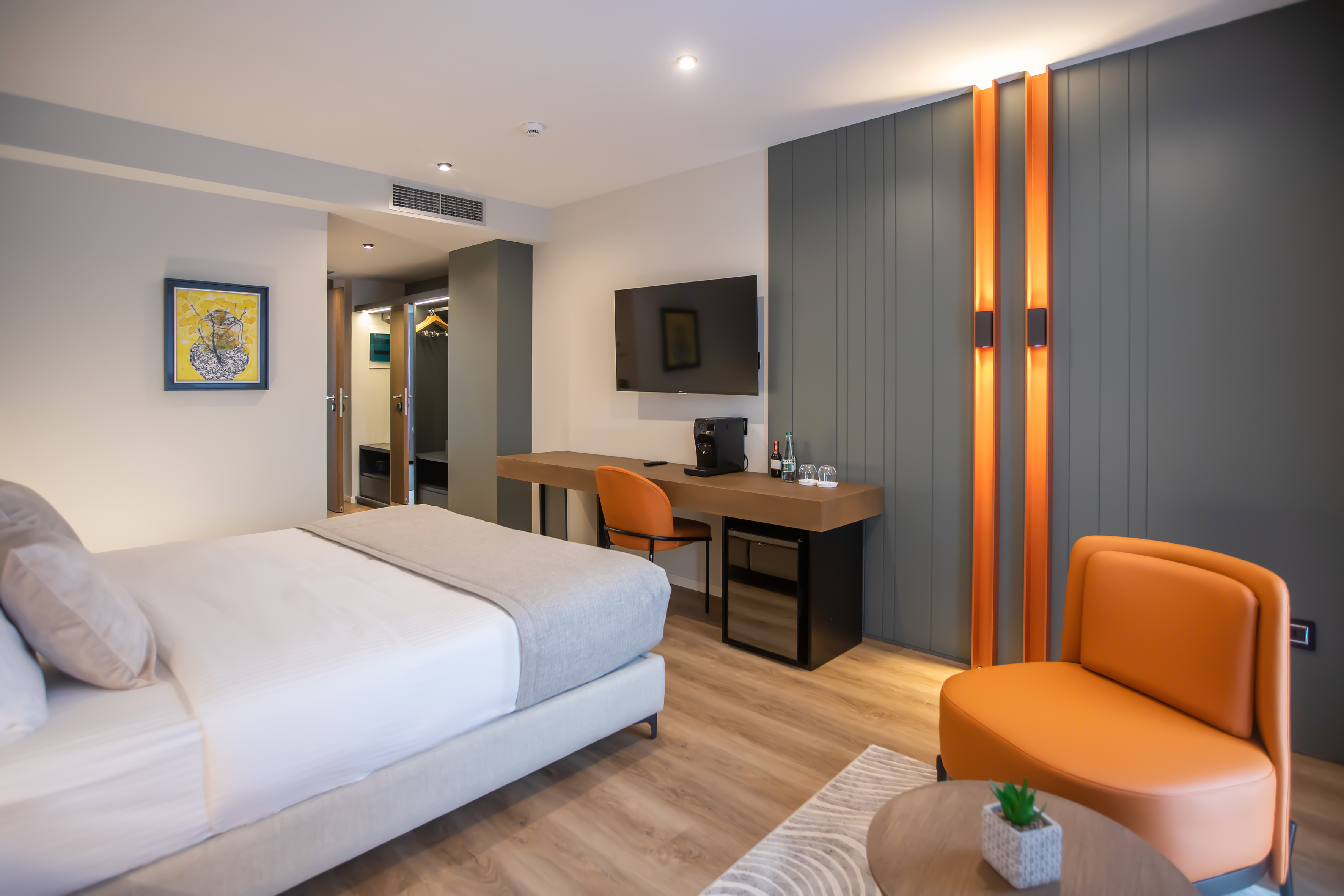 Modern hotel room with bed, orange chairs, desk, TV, and contemporary design.