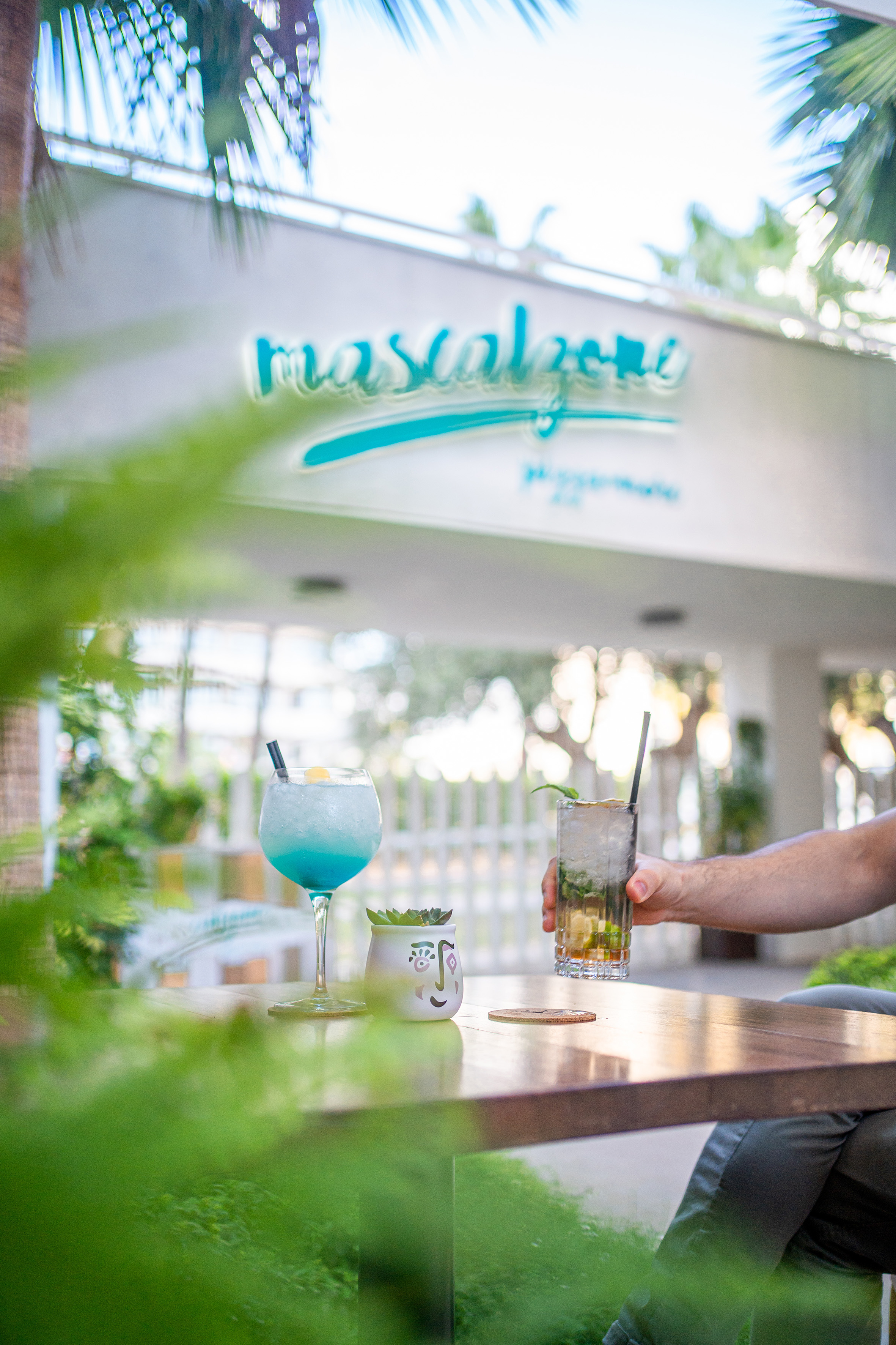 Mascalzone patio: blue cocktail, mojito, and a hand holding a drink on table.