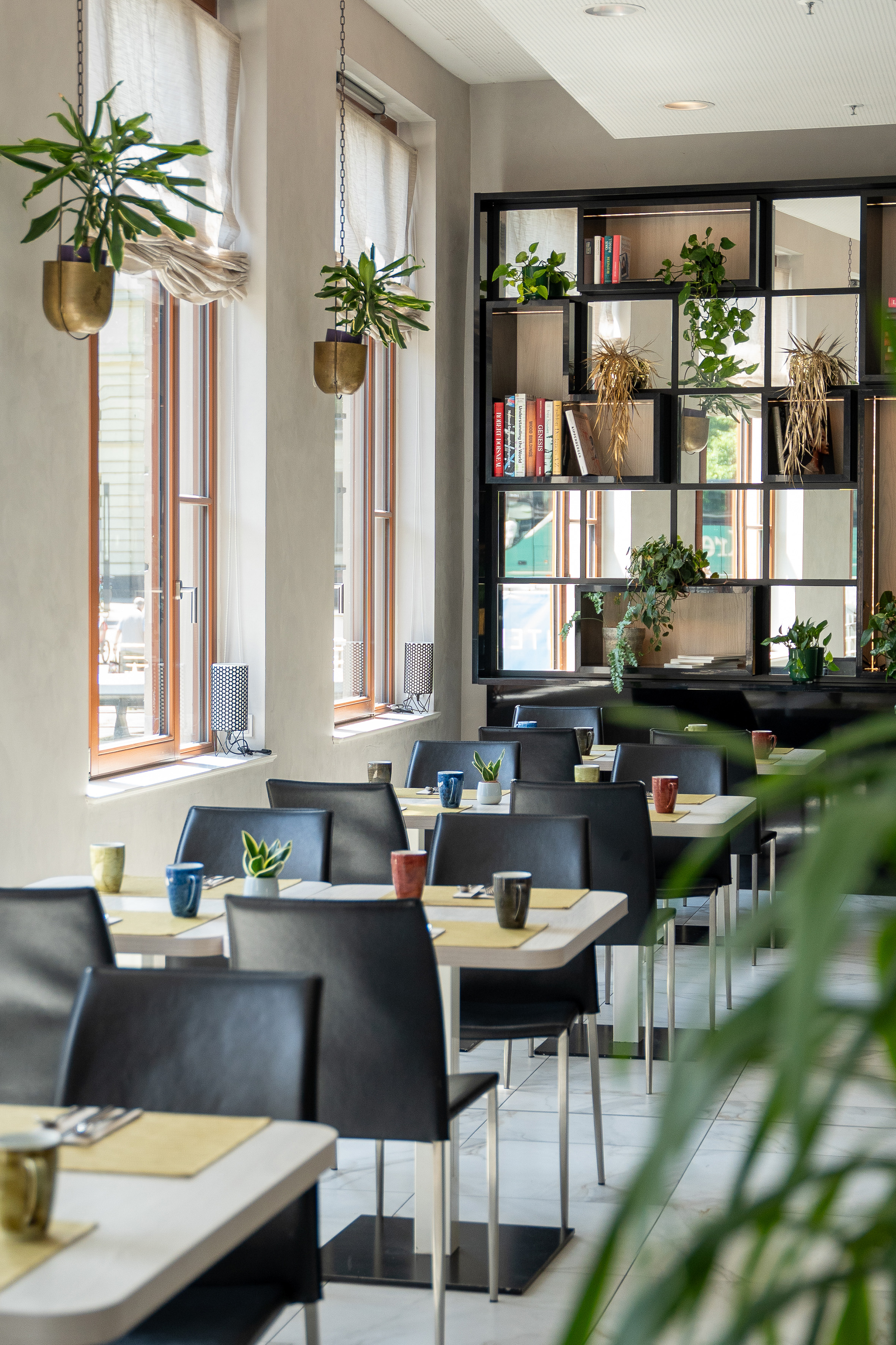 Modern cafe dining area with tables, black chairs, green plants, and a bookshelf.