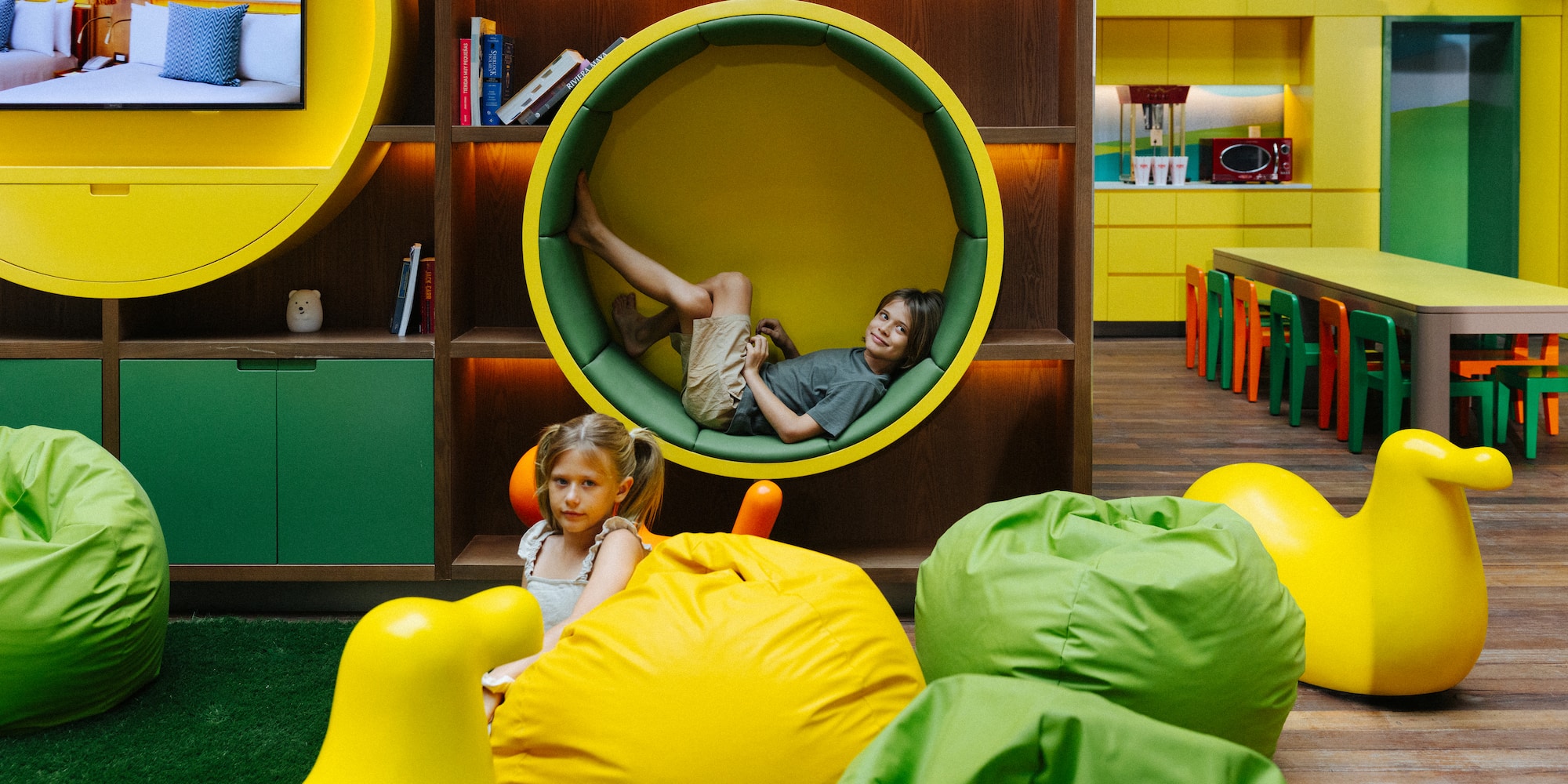 Boy in circular reading nook, girl on yellow bean bag in modern kids play area.