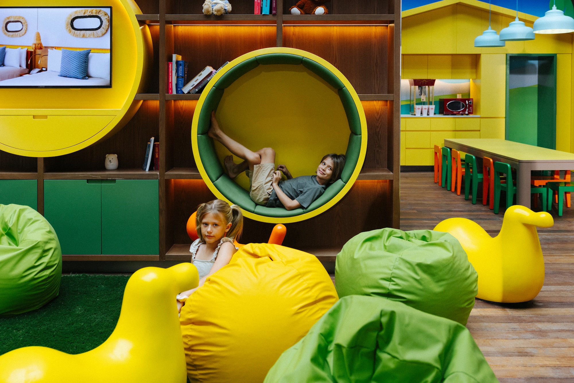 Boy in circular reading nook, girl on yellow bean bag in modern kids play area.