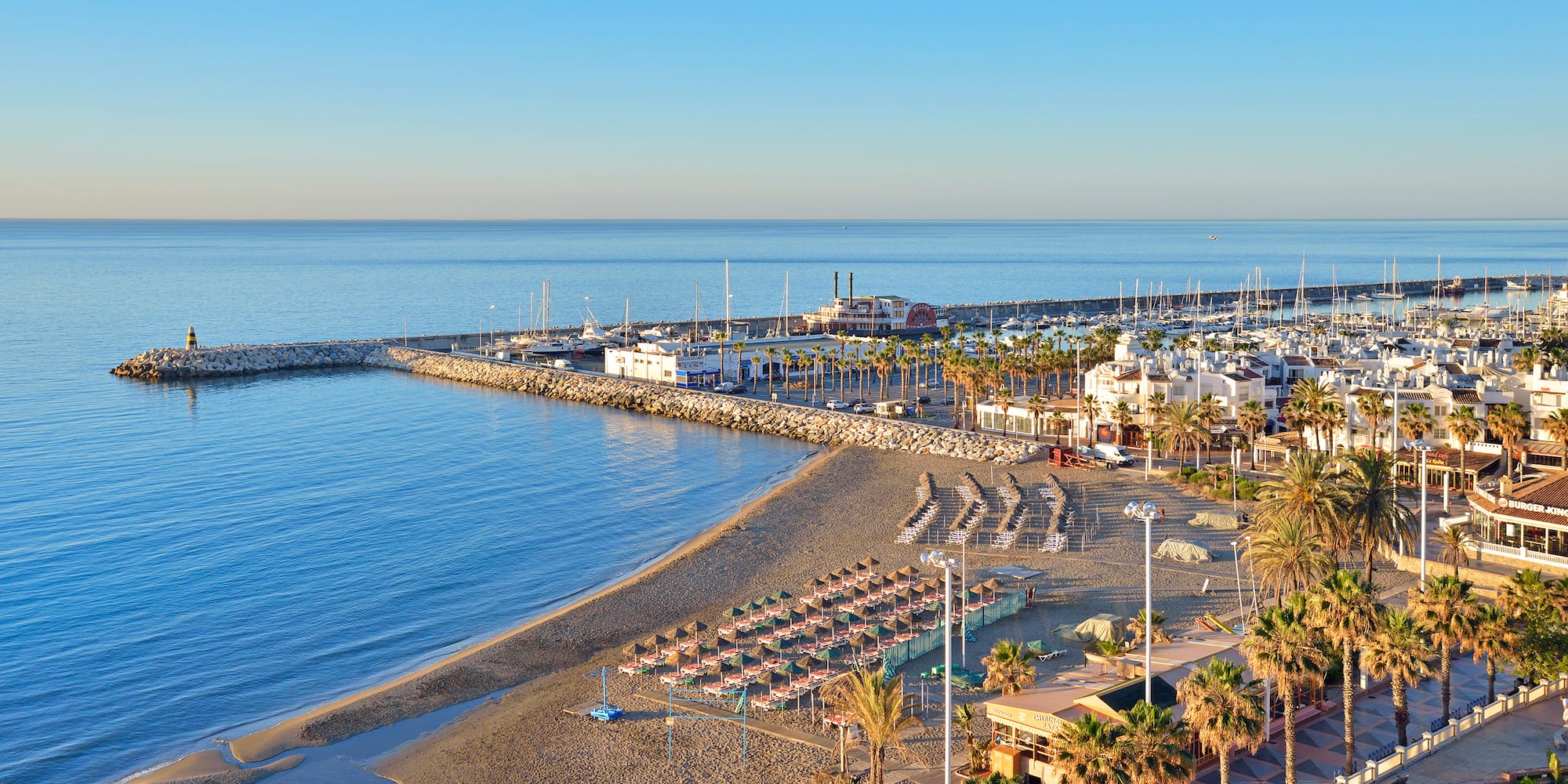 Scenic resort beach with marina, yachts, stone pier, and palm trees.