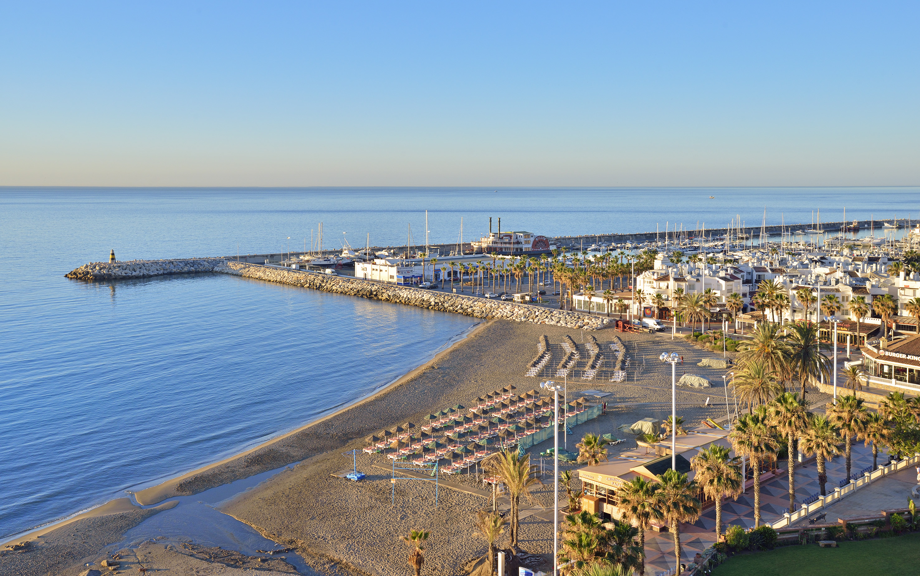 Scenic resort beach with marina, yachts, stone pier, and palm trees.