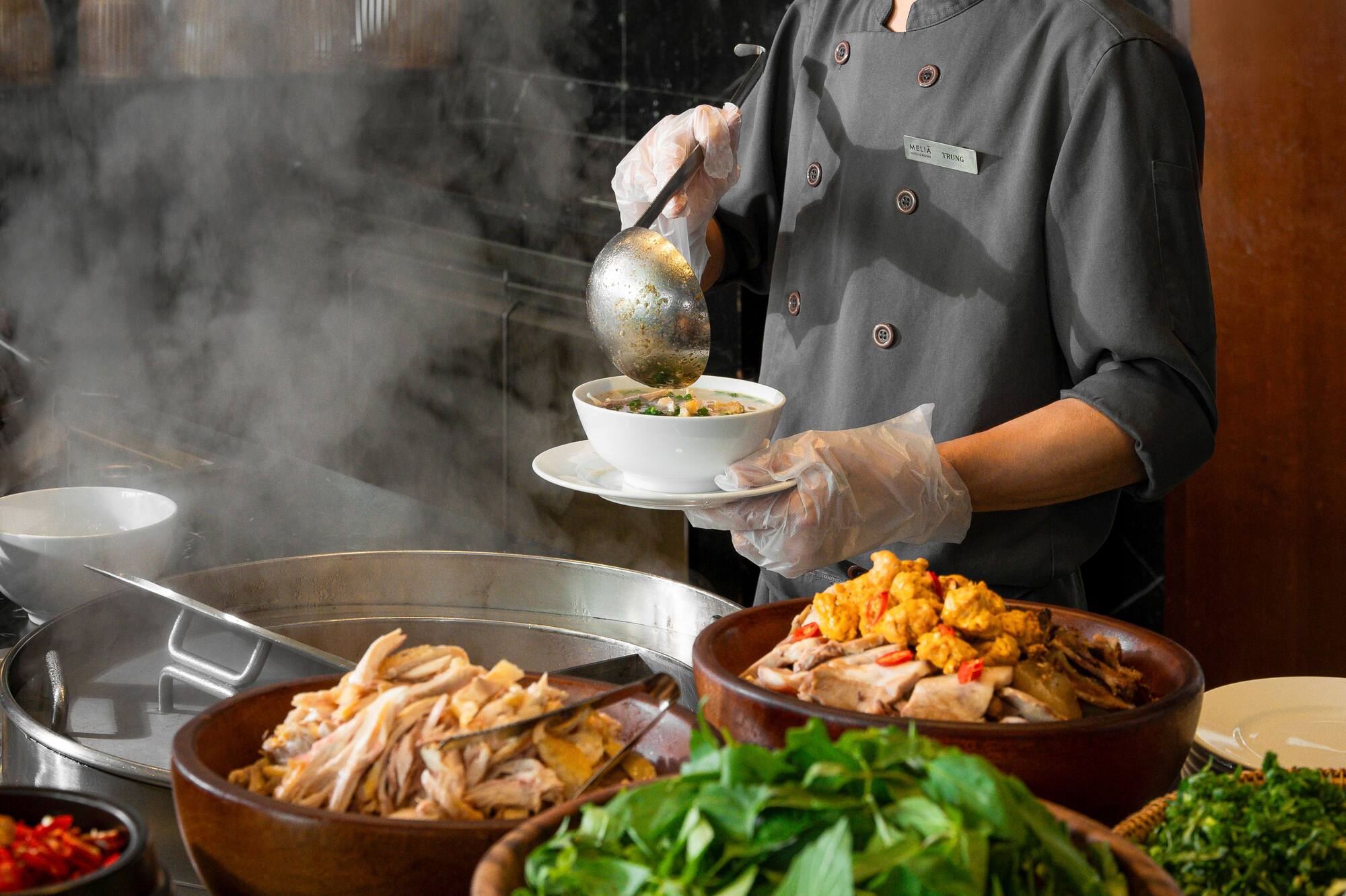 Chef serving hot noodle soup at a buffet with fresh chicken and herbs.