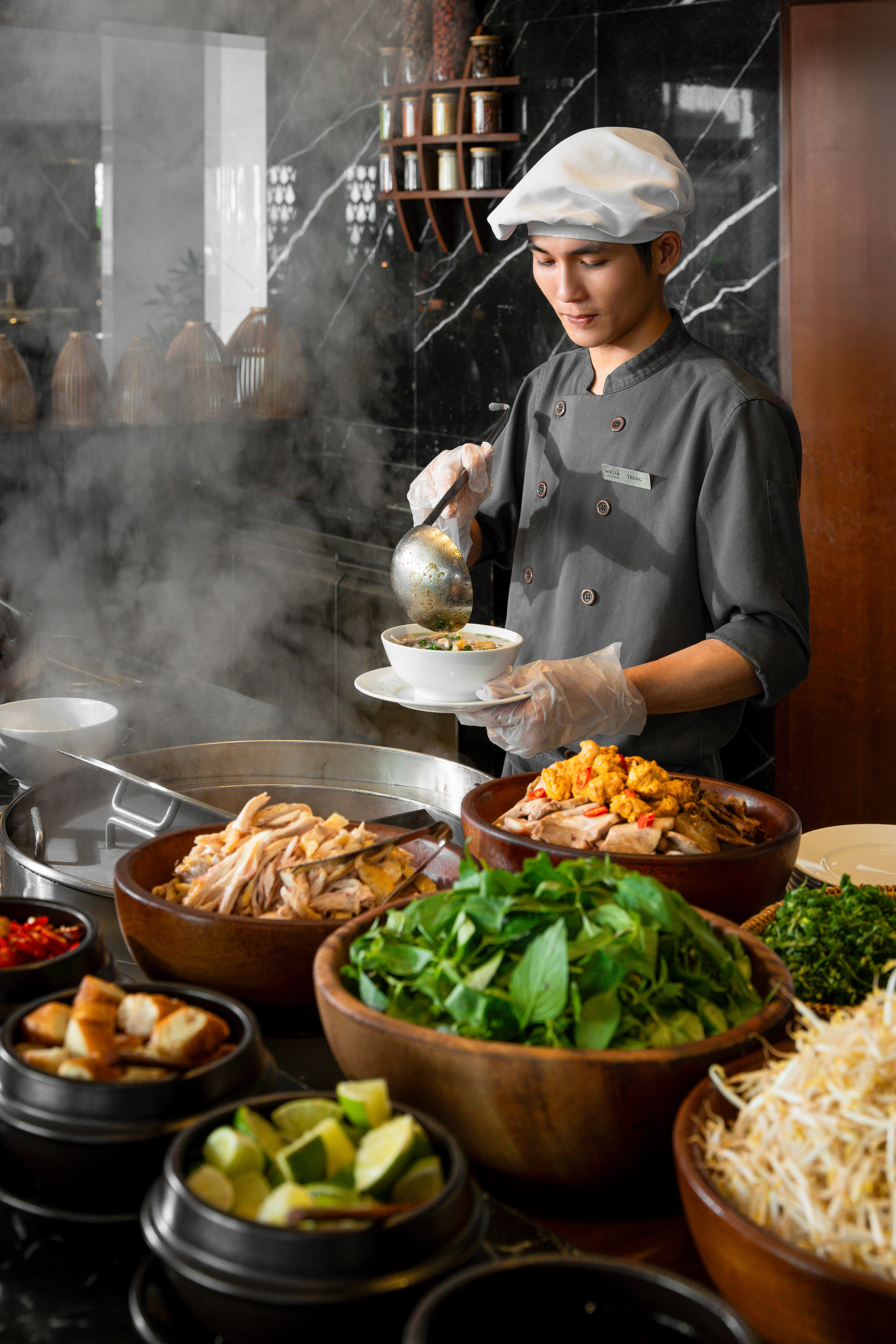 Chef serving hot noodle soup at a buffet with fresh chicken and herbs.