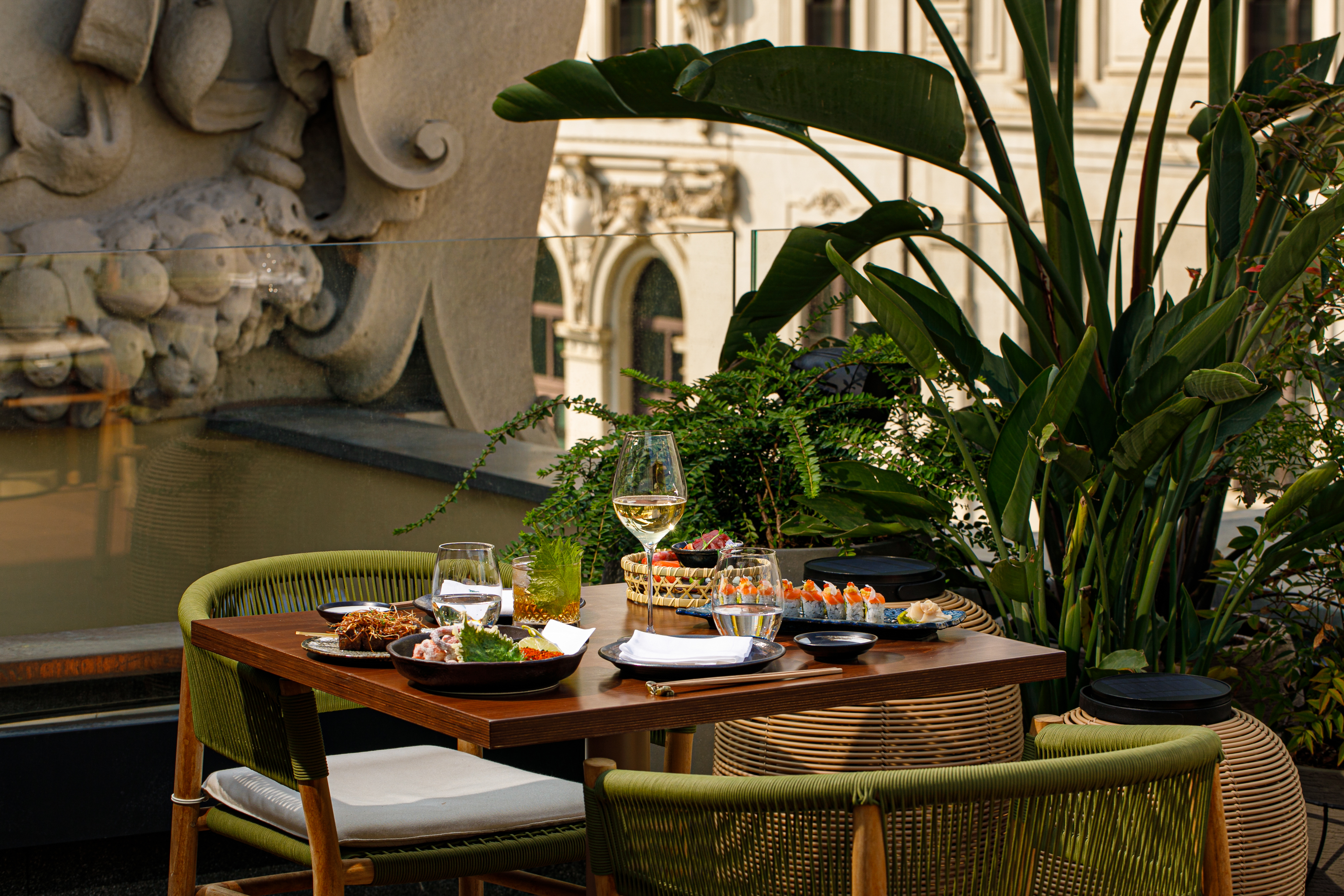 Rooftop restaurant table with sushi, wine, and lush plants, city view.